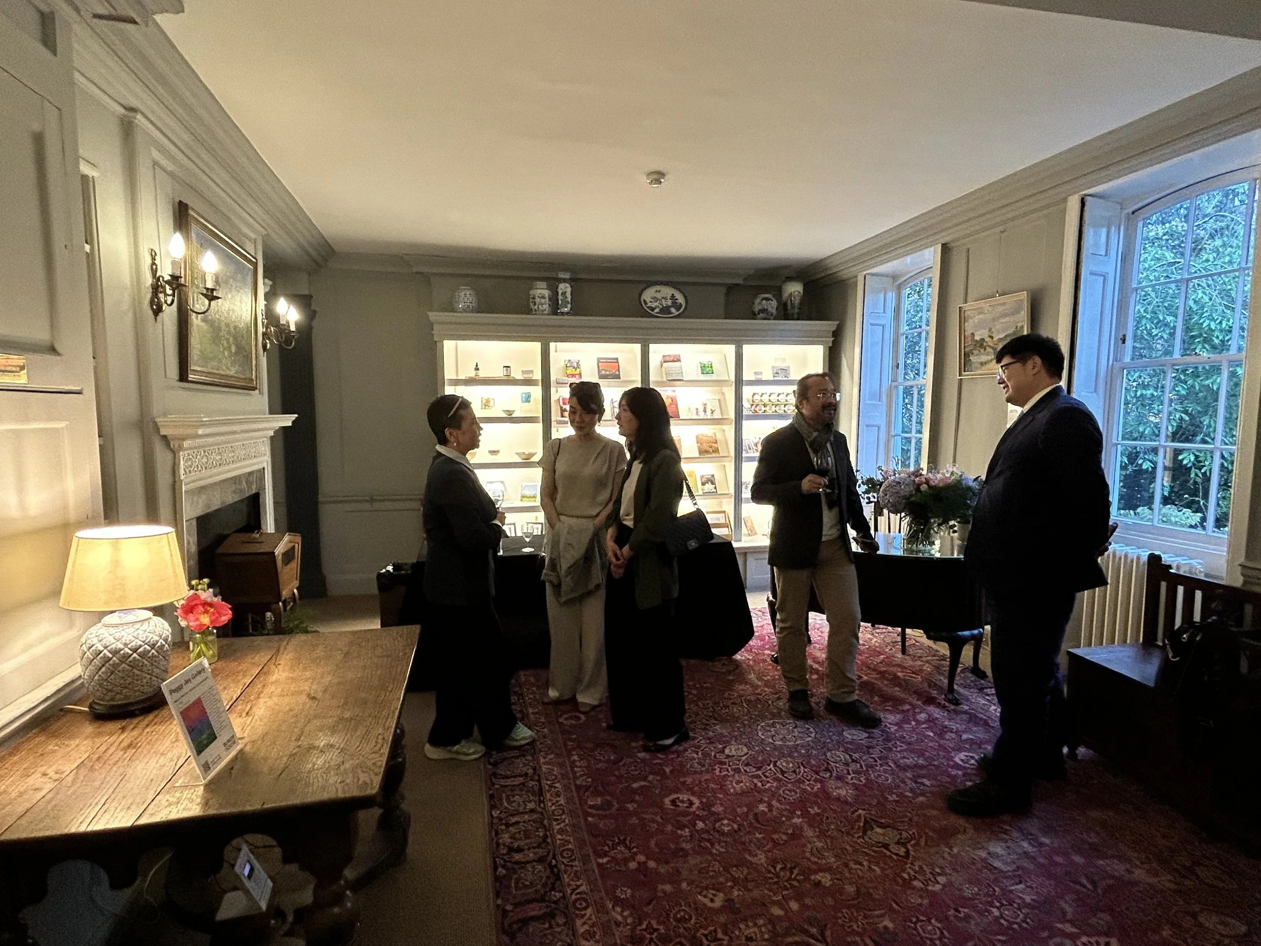 Group of five people talking in a cozy, well-decorated room with large windows, a fireplace, a wooden table, and display shelves filled with books and decorative items.