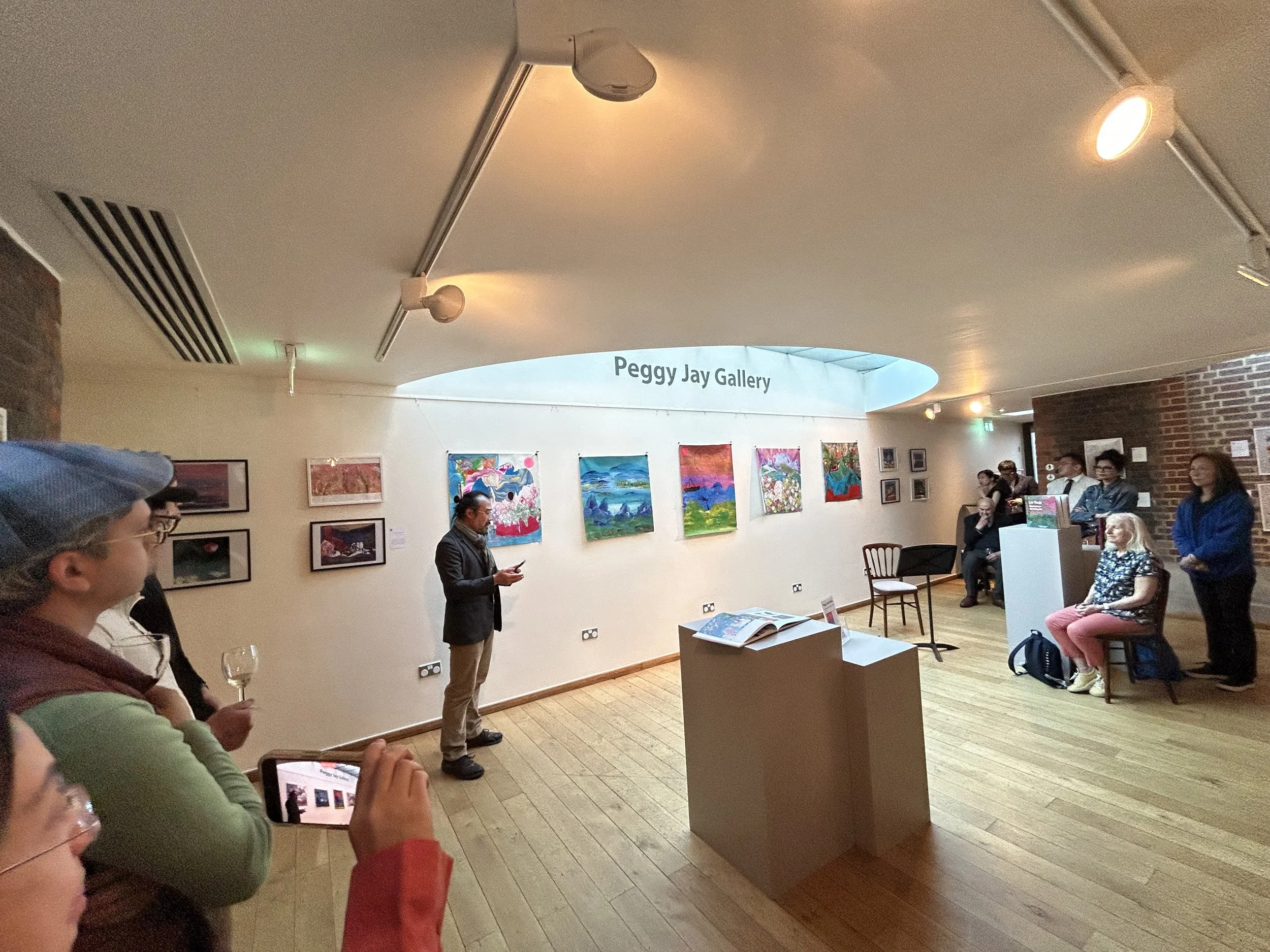 People attending an art gallery exhibit, with paintings displayed on the walls and a sign reading 'Peggy Jay Gallery.' Some visitors are seated, others are standing and engaging with the artwork, and one person is using a tablet.