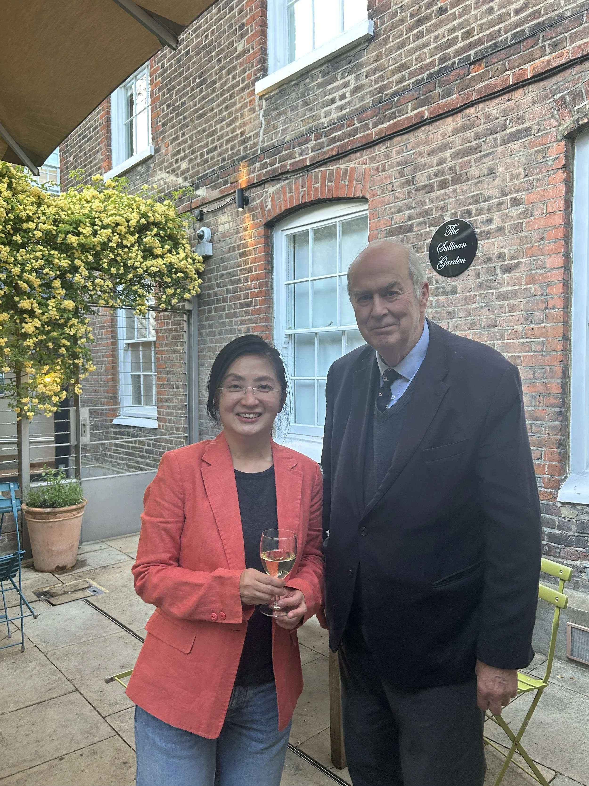 A woman in a coral blazer and jeans holding a glass of white wine, standing next to an older man in a dark suit outside on a patio, with a brick building and green plants in the background.