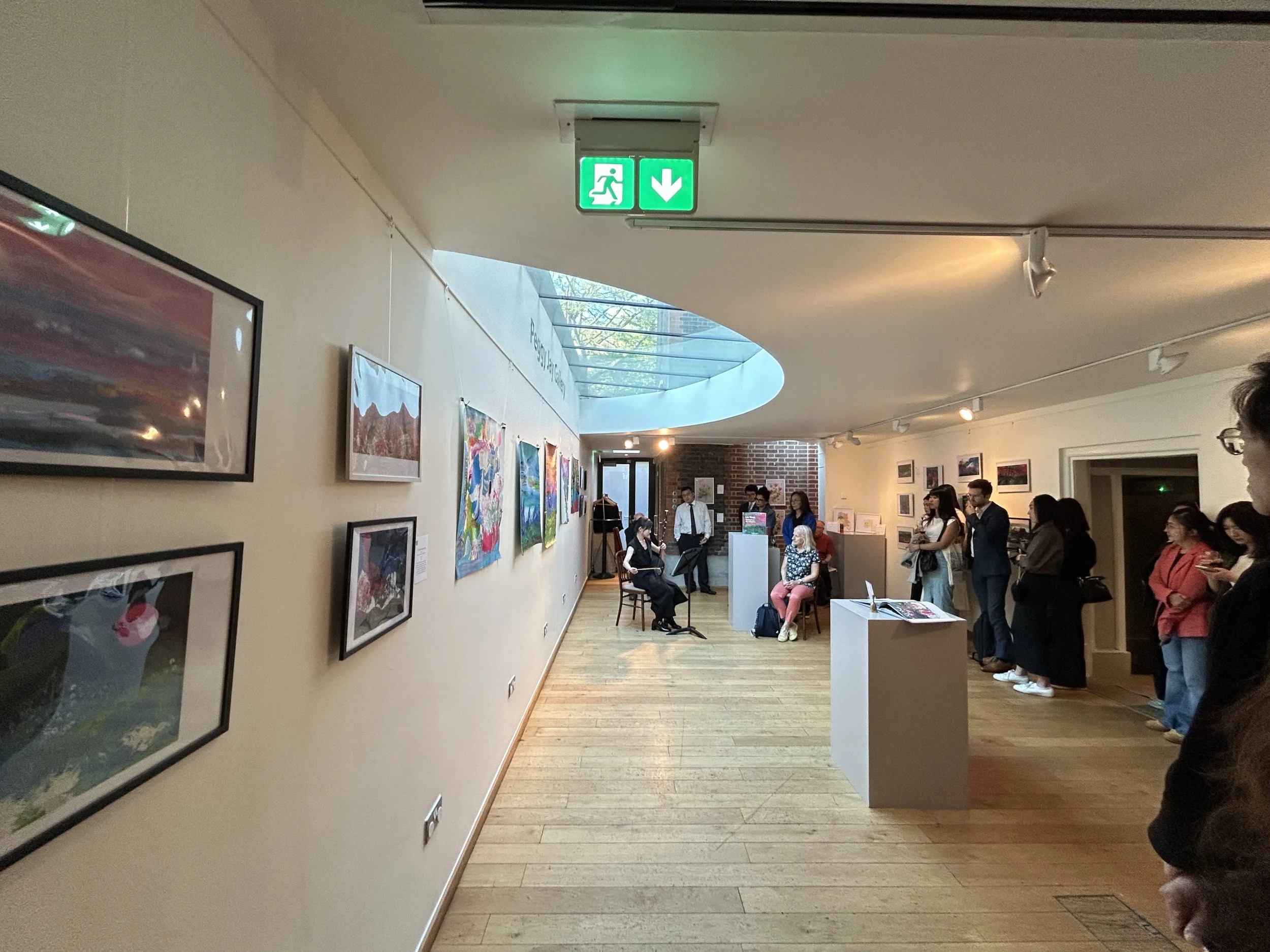 An indoor art gallery with visitors standing and sitting, artwork displayed on the walls, and a bright skylight overhead.
