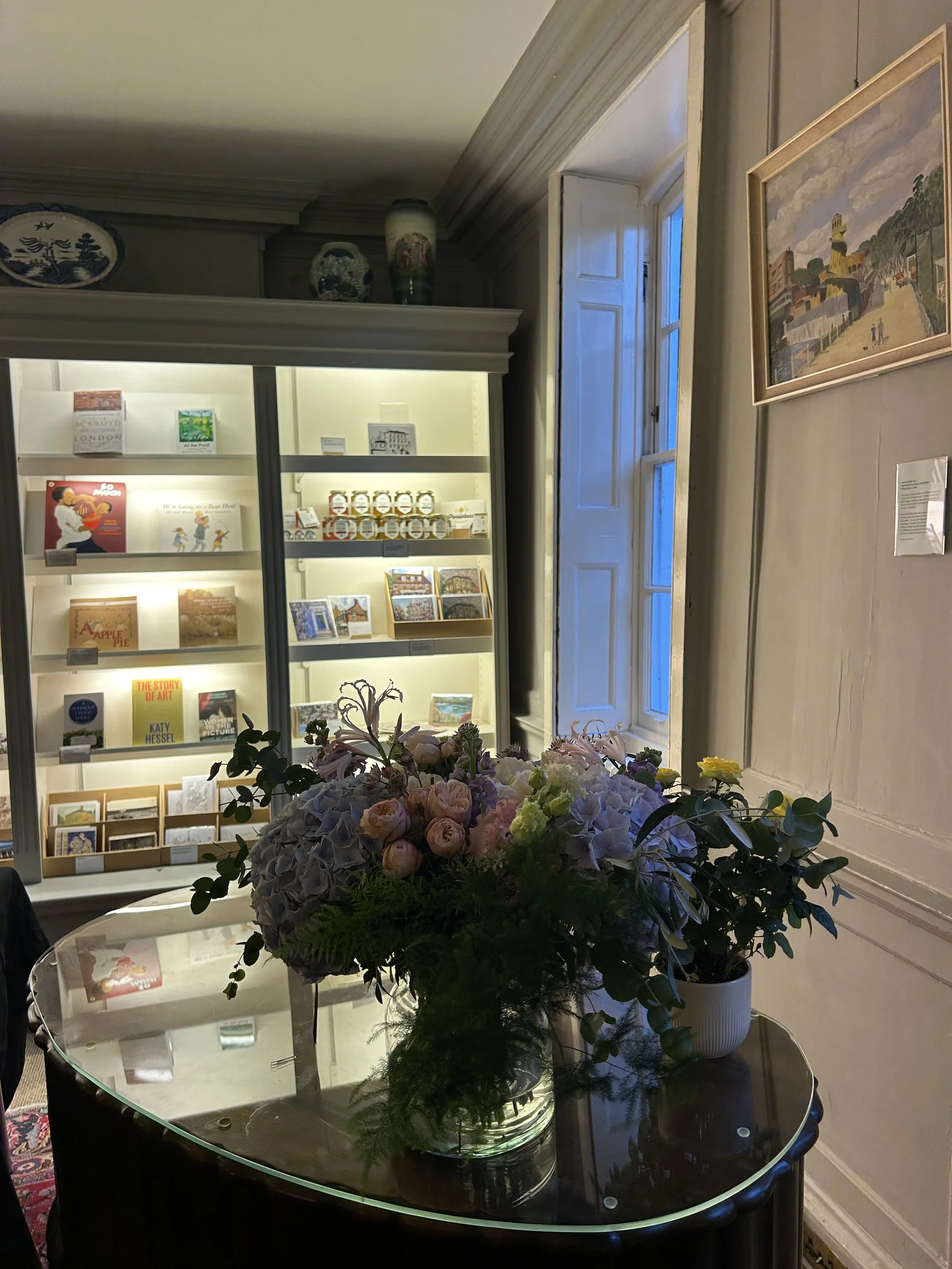 A floral arrangement featuring pink, white, and purple flowers on a glass table in front of a display cabinet filled with books and jars, with a window and painting on the wall in the background.