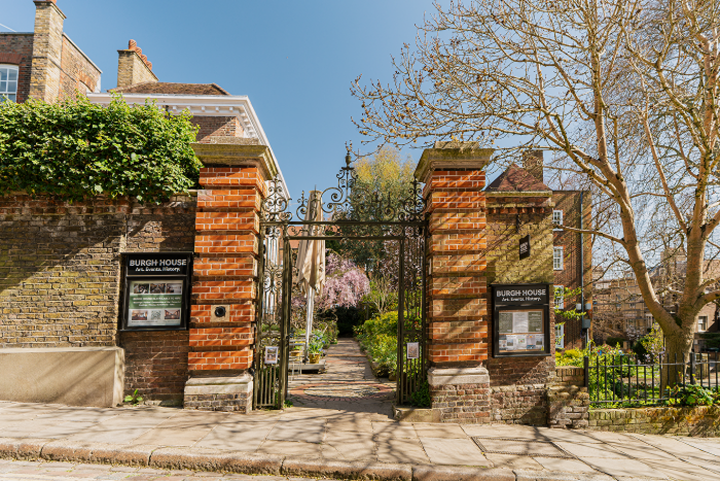Entrance to Burgh House with iron gate and brick pillars, view into a garden with pathways and flowering trees, informational signs on either side of the gate, clear blue sky overhead.