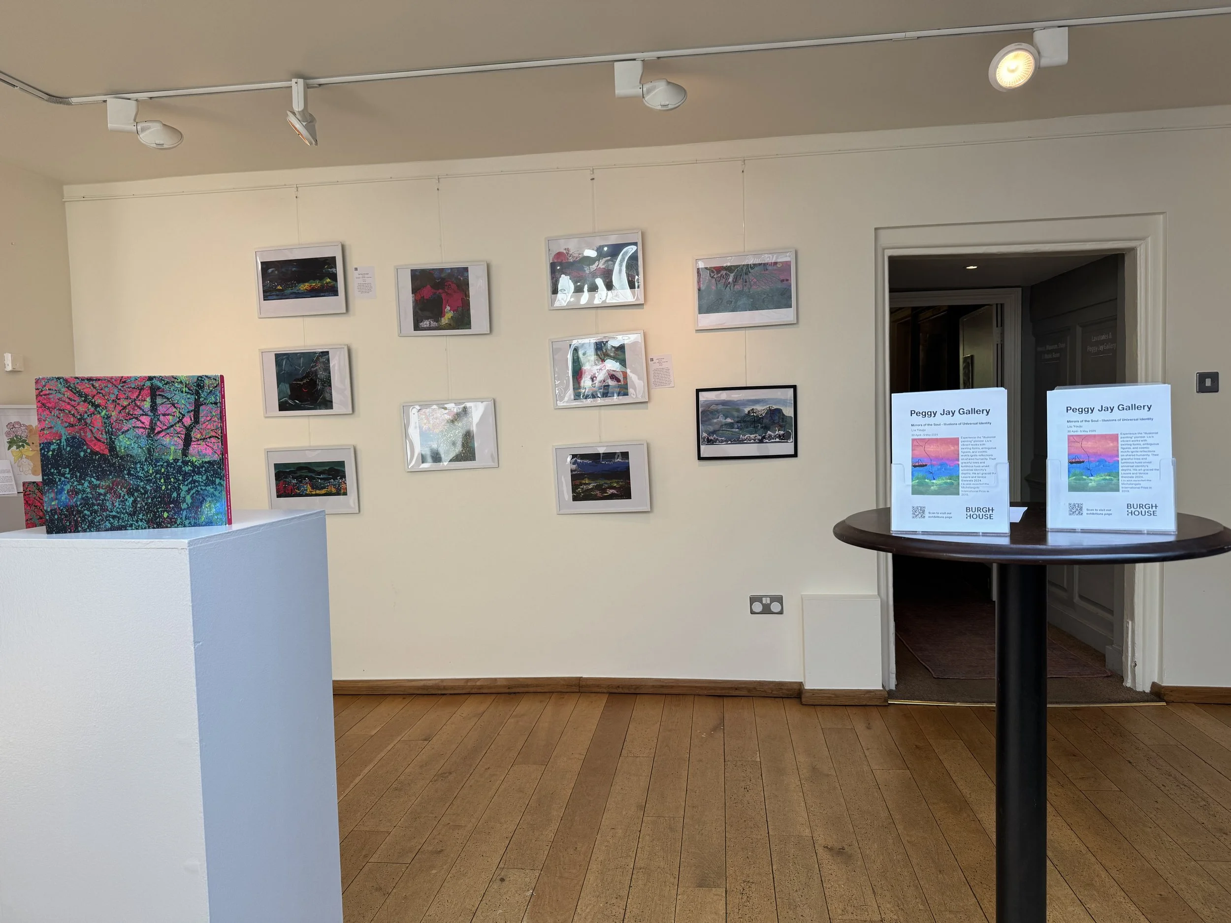 An art gallery display with framed artworks on the wall and informational signs on a round table in the foreground. The gallery has light-colored walls and a wooden floor.