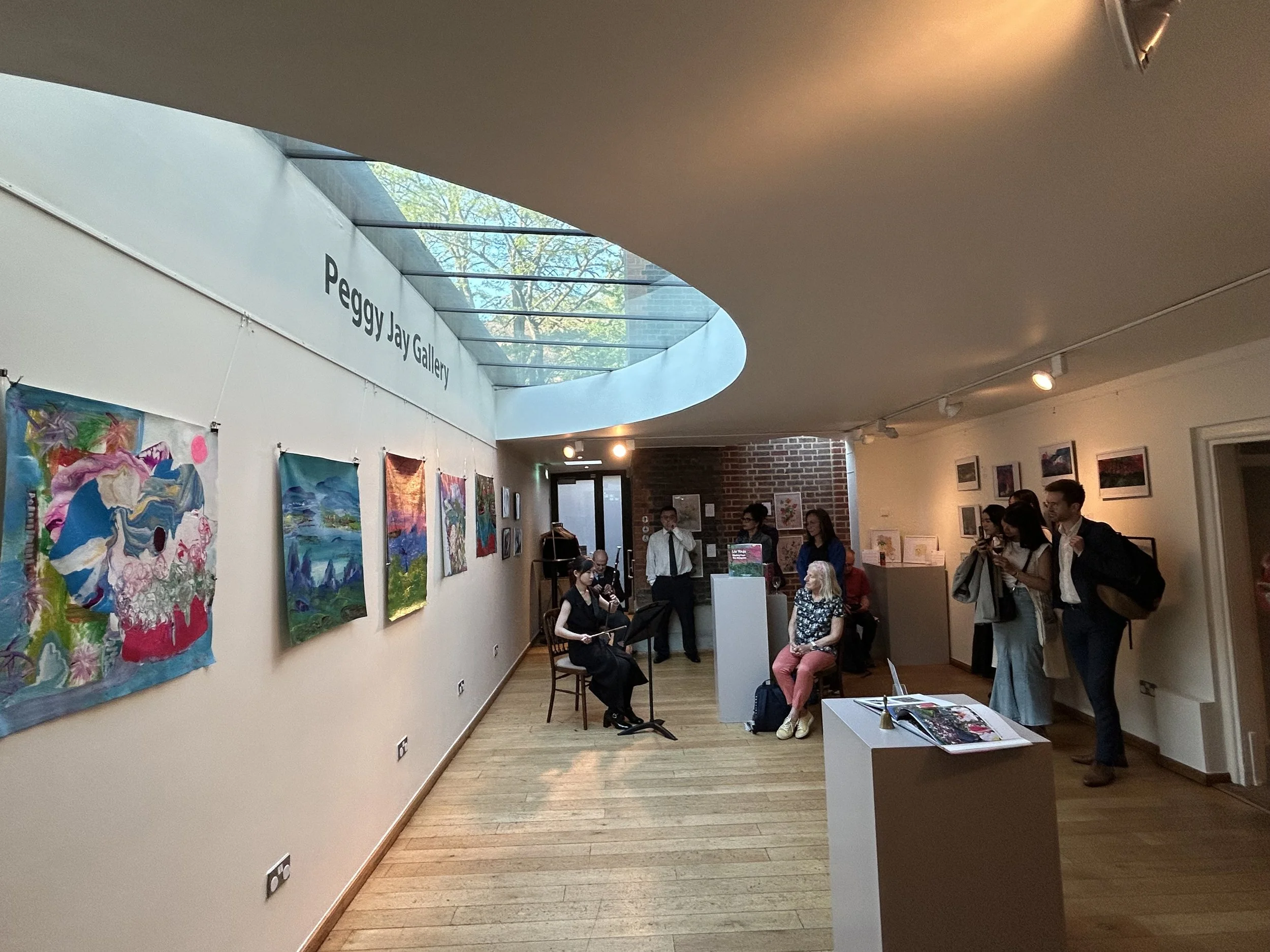 Art gallery exhibition with colorful paintings on the wall and people viewing and discussing the artwork inside a modern building with a glass ceiling.