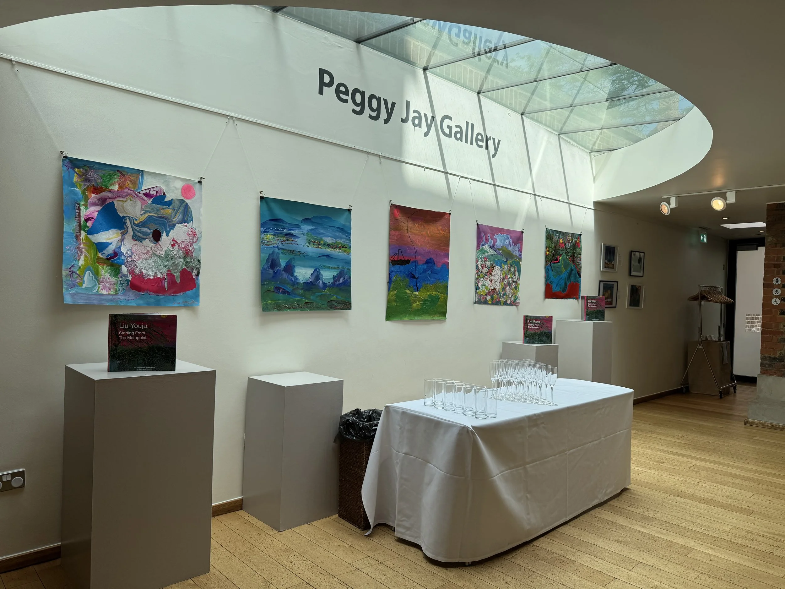Interior of an art gallery with paintings hanging on a white wall. The gallery is named Peggy Jay Gallery, as indicated by large text on the wall near a glass ceiling. There are tall white pedestals with books and a table with glasses beneath a white