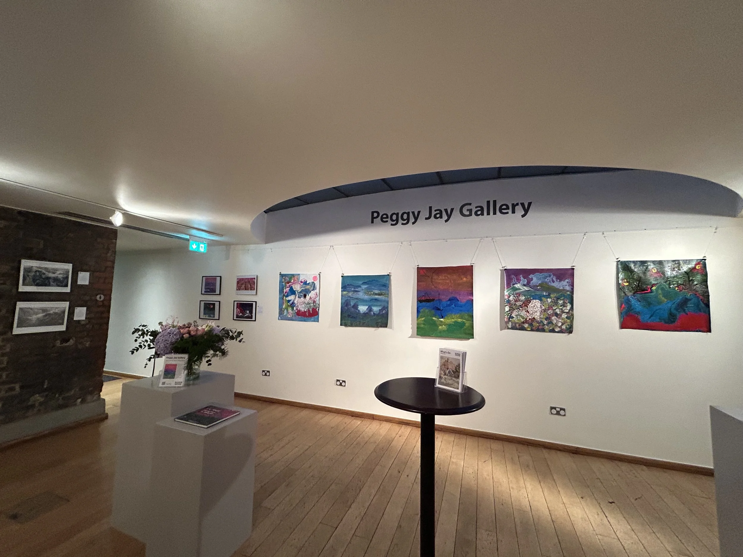 An art gallery with colorful paintings displayed on white walls, featuring a sign that reads 'Peggy Jay Gallery' and a flower arrangement on a pedestal.