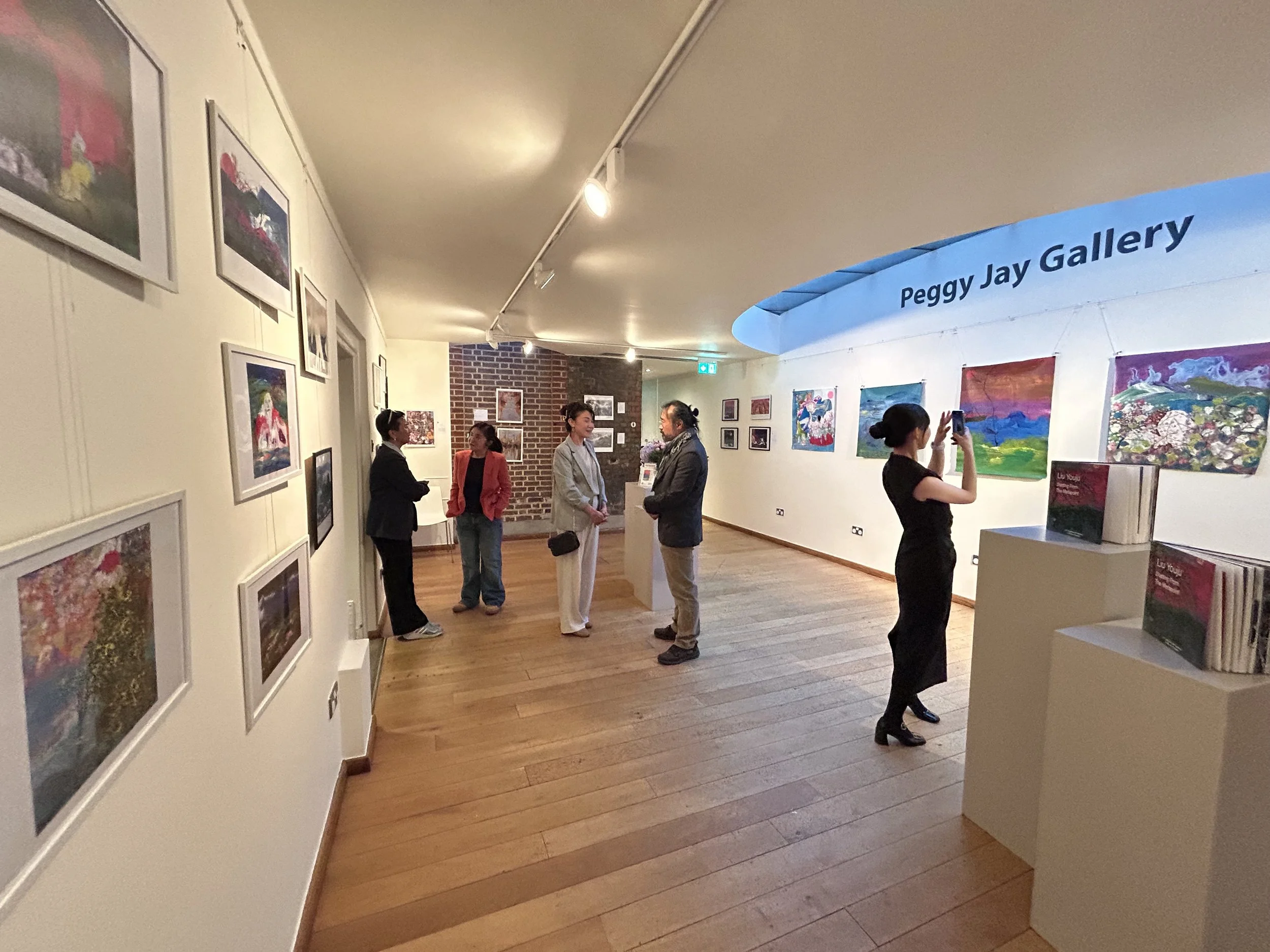 People viewing artwork inside an art gallery with a sign reading 'Peggy Jay Gallery' on the wall.