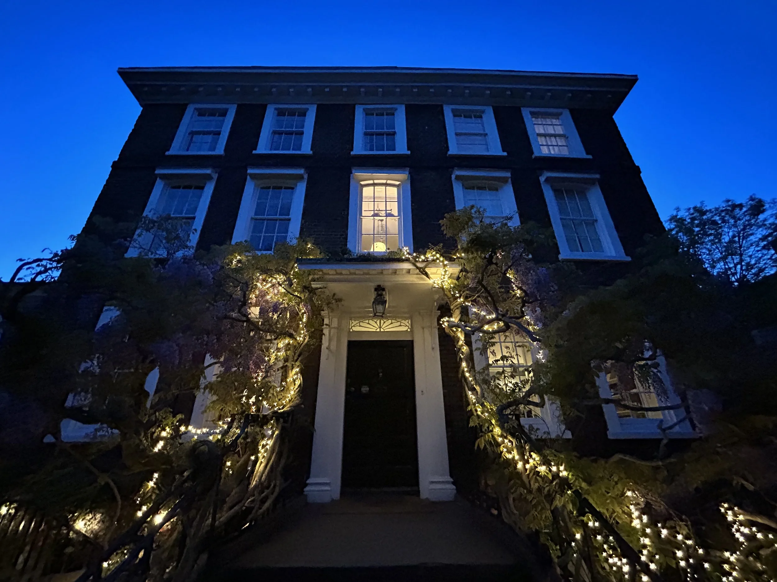 A dark brick multi-story house with lit windows at night, decorated with string lights on the surrounding trees.