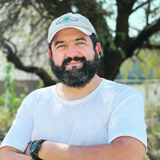 Hombre con barba, camiseta blanca, gorra blanca, de fondo un árbol y un día soleado.