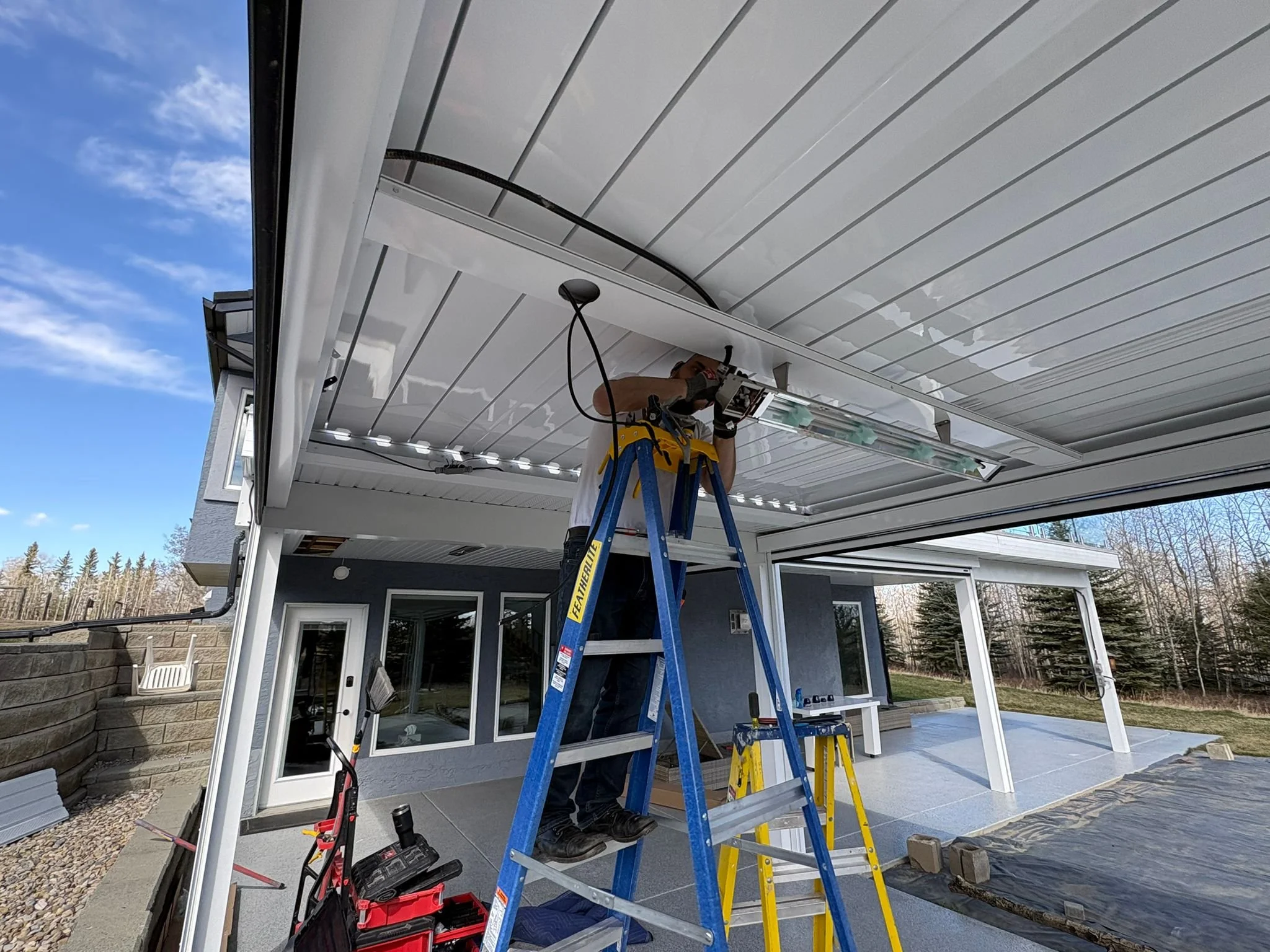 Person standing on a blue ladder installing or repairing a light fixture on a white ceiling under a covered patio.