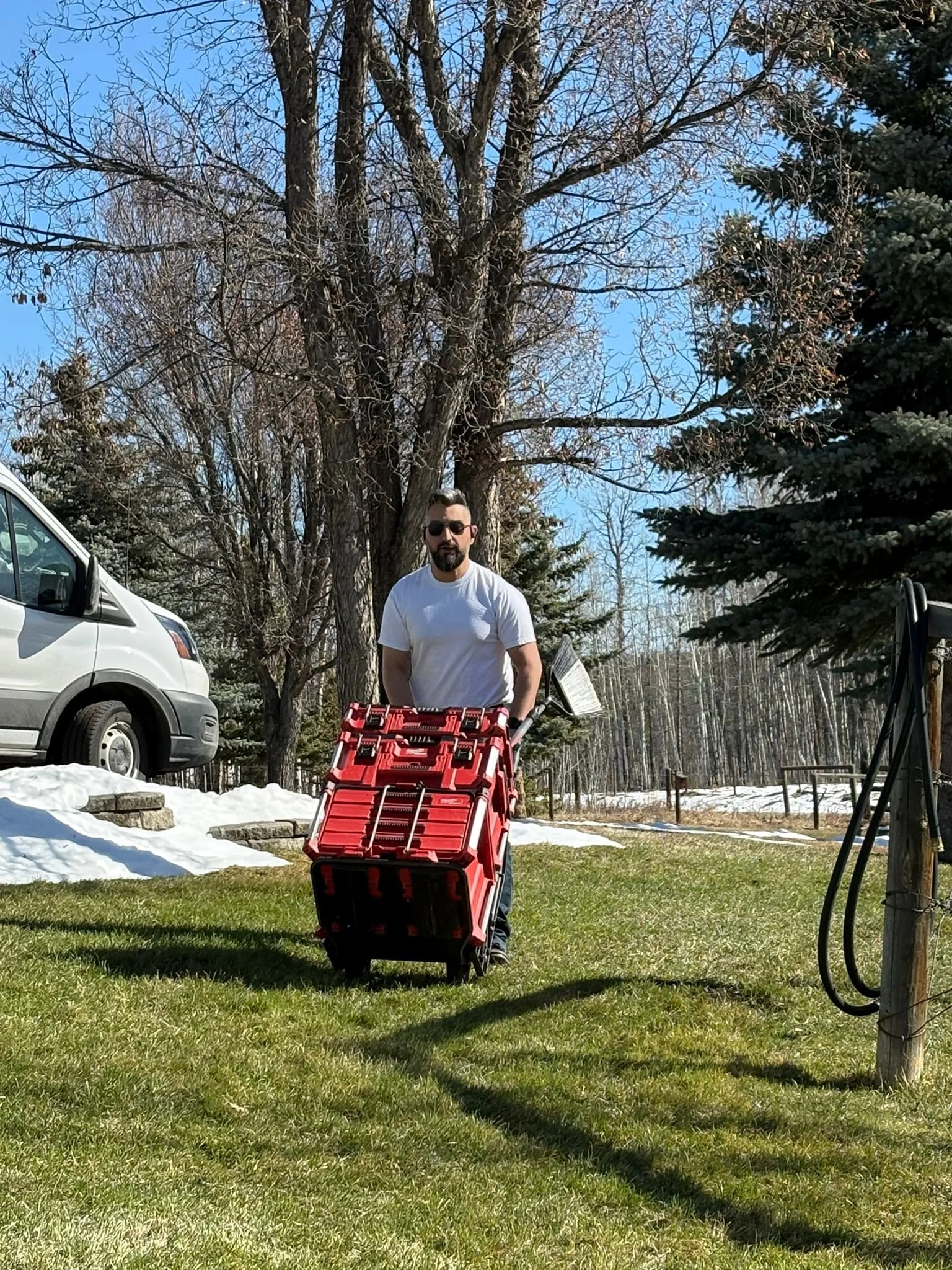 A man with sunglasses, a white t-shirt, and dark hair in a top knot, standing outdoors on grass with snow patches, holding a red and black toolbox or equipment case. In the background, there are large trees, a white van, and a wooded area. The sky is clear and blue.