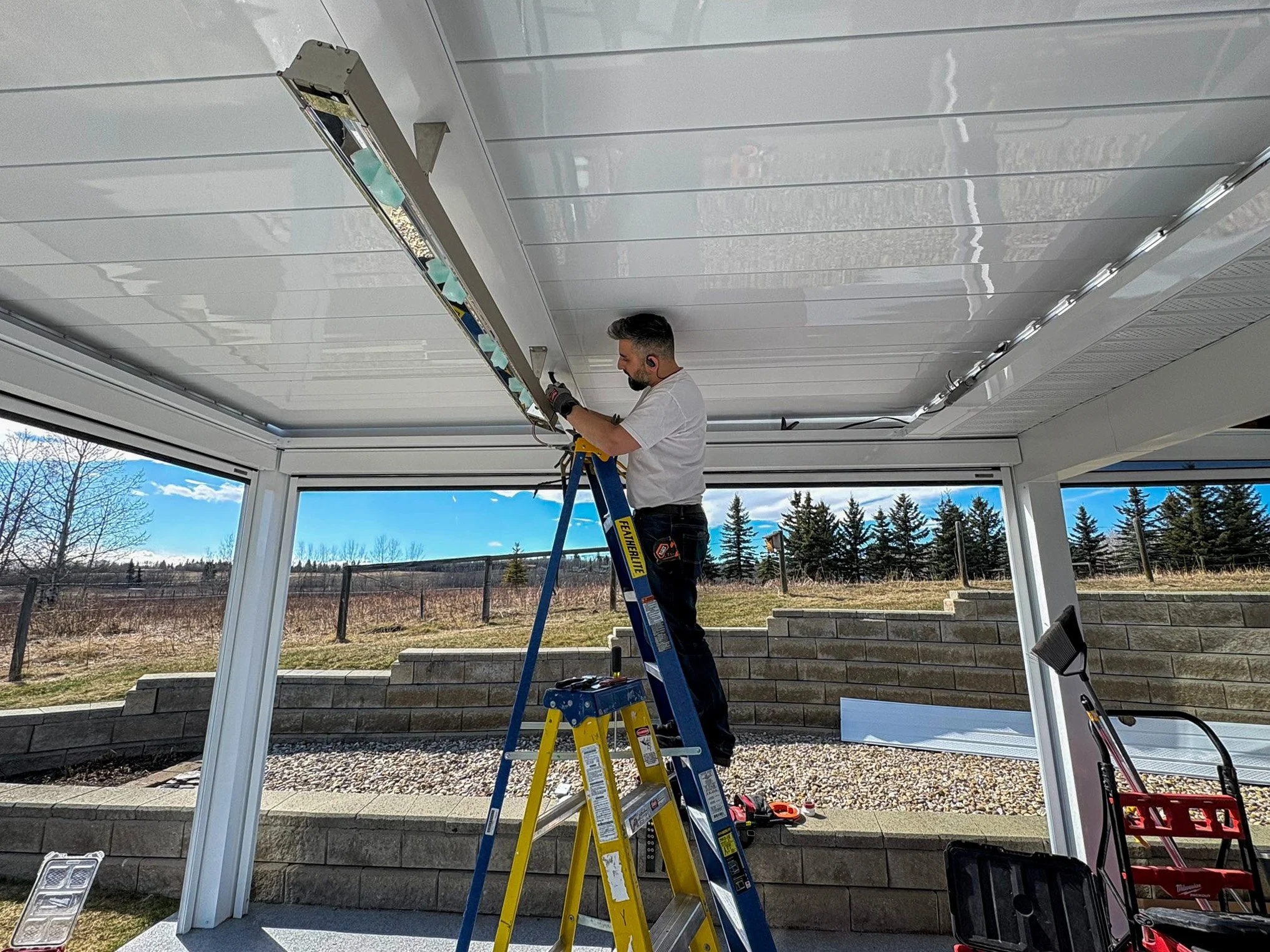 A man wearing a white t-shirt and black gloves stands on a ladder, working on a ceiling light fixture or electrical installation in an enclosed patio or sunroom. Outside views include blue sky, some trees, and a grassy area.