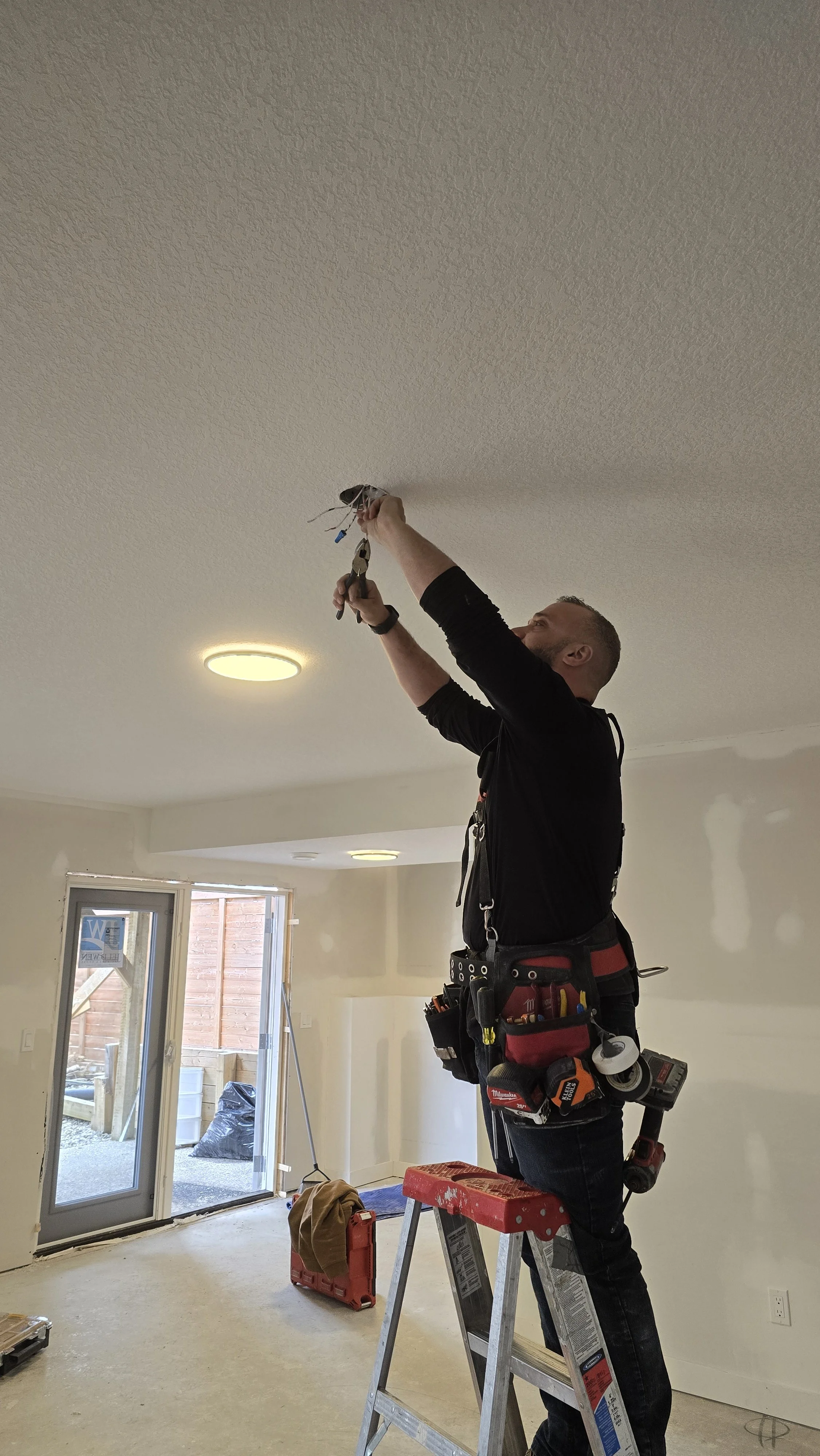 A man on a ladder working on electrical wiring in a room under a textured ceiling.
