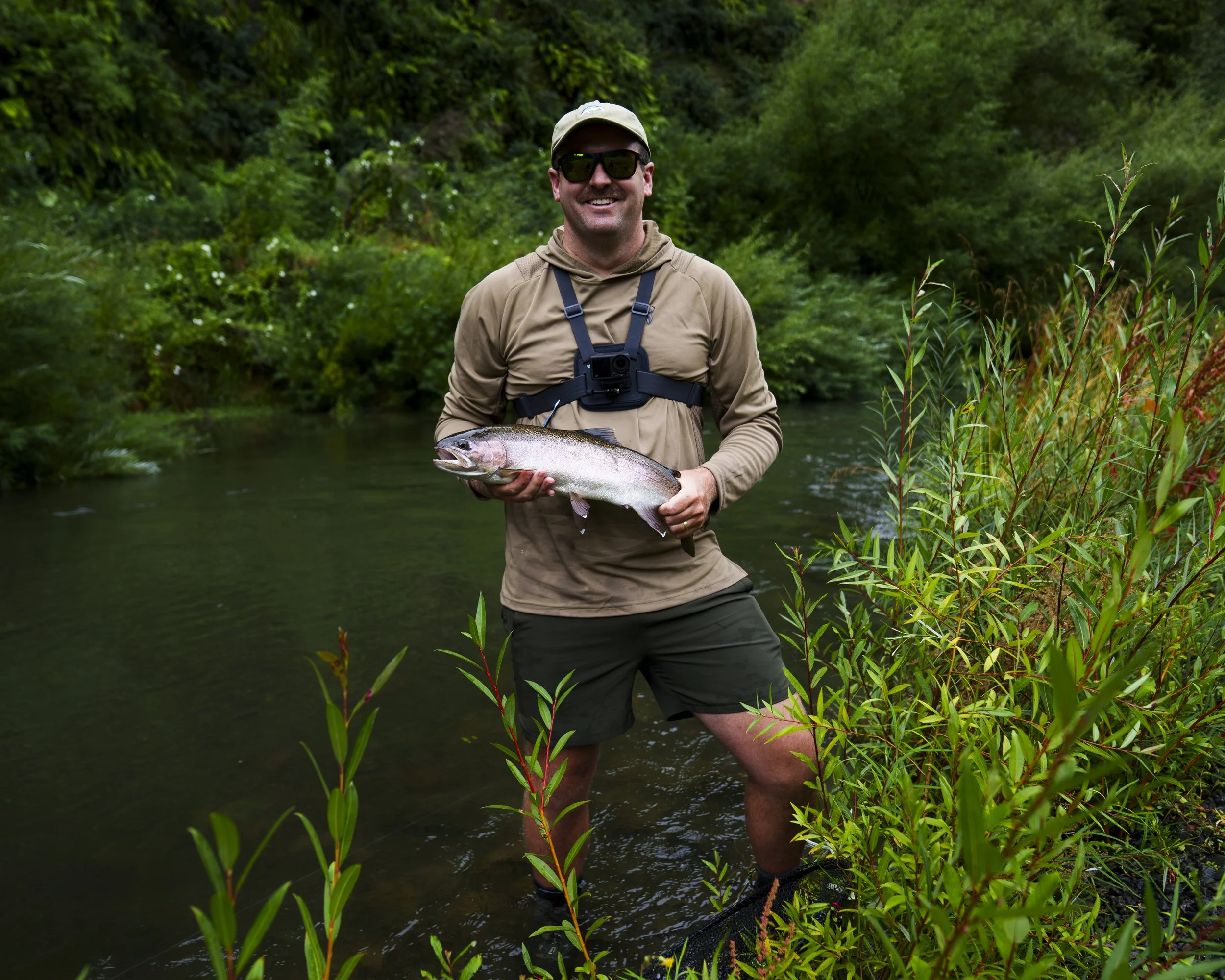 A man standing in a river, holding a large fish, surrounded by lush green vegetation.