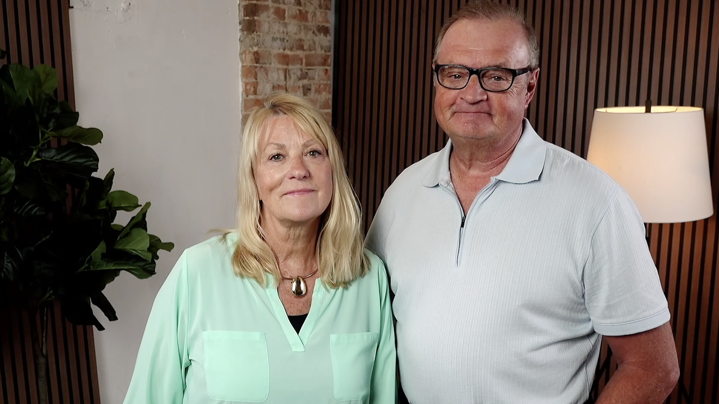 Two older adults, a woman with blonde hair and a man with glasses, standing side by side indoors. The woman is wearing a mint green top with a large pendant necklace, and the man is wearing a light grey polo shirt. There is a plant on the left, a brick wall, wooden panels, and a lamp in the background.
