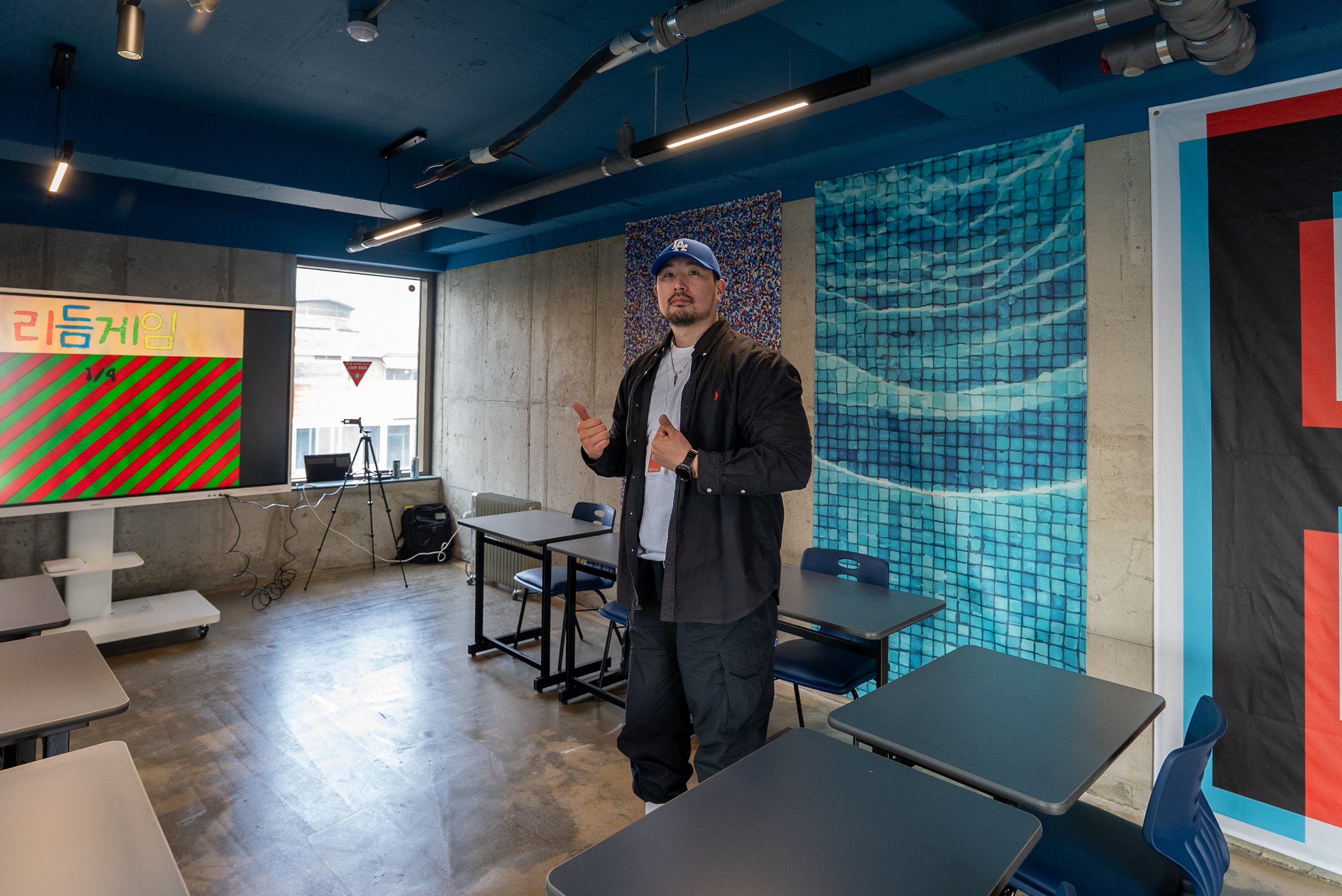 Man standing in a classroom or office with tables and chairs, wearing a black jacket, white shirt, and a blue LA Dodgers cap, giving a thumbs-up.