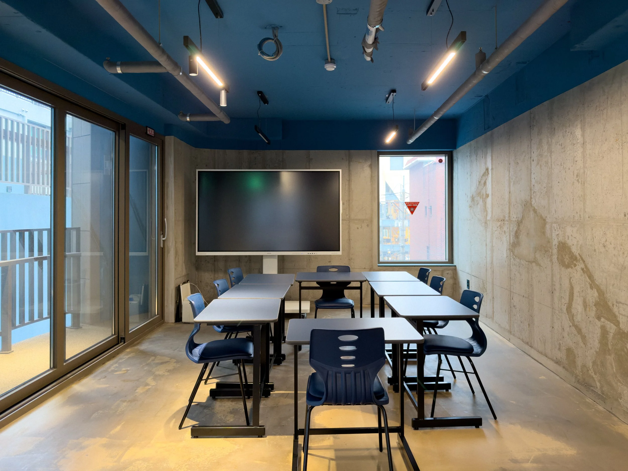 Modern conference room with tables and chairs, large TV screen, glass doors, concrete walls, and window showing city buildings.