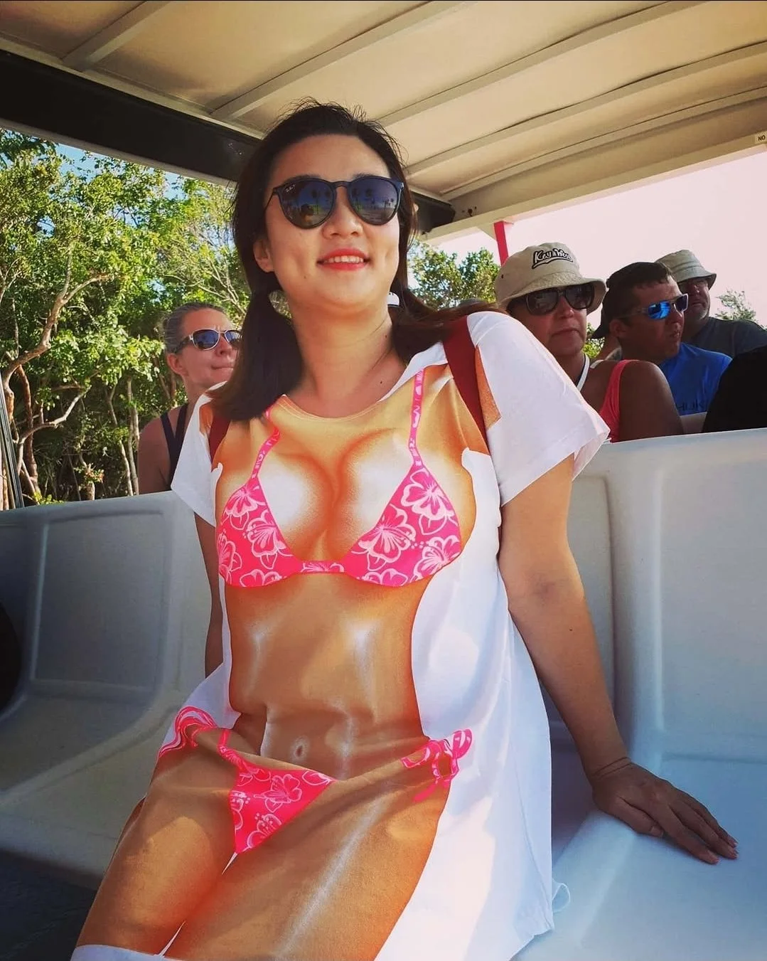 Woman wearing sunglasses and a T-shirt with a printed image of a bikini-clad woman on it, sitting on a train with other passengers in the background, some wearing hats and sunglasses, with trees visible outside.