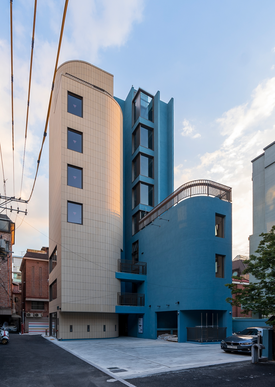 Modern multi-story building with blue and beige exterior, located in an urban area under a partly cloudy sky.