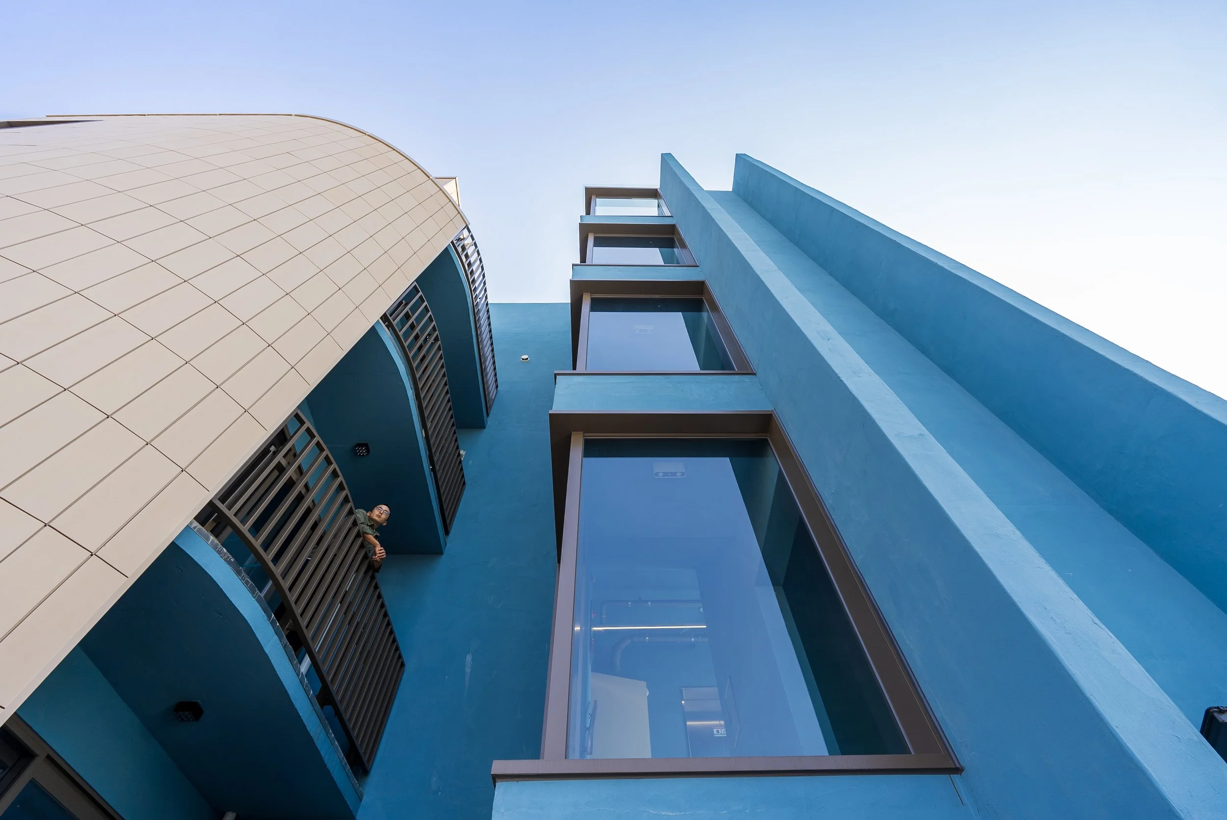 Low-angle view of modern blue building with large glass windows and balcony, against a clear sky. A woman is on a small balcony.
