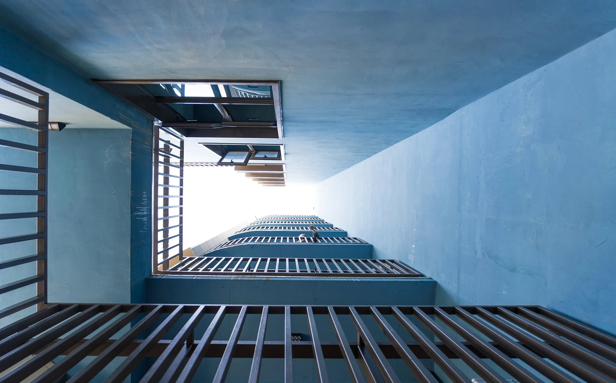 View of a modern apartment building from the ground level looking upward, with prominent blue exterior walls, metal railings, and windows against a bright sky.