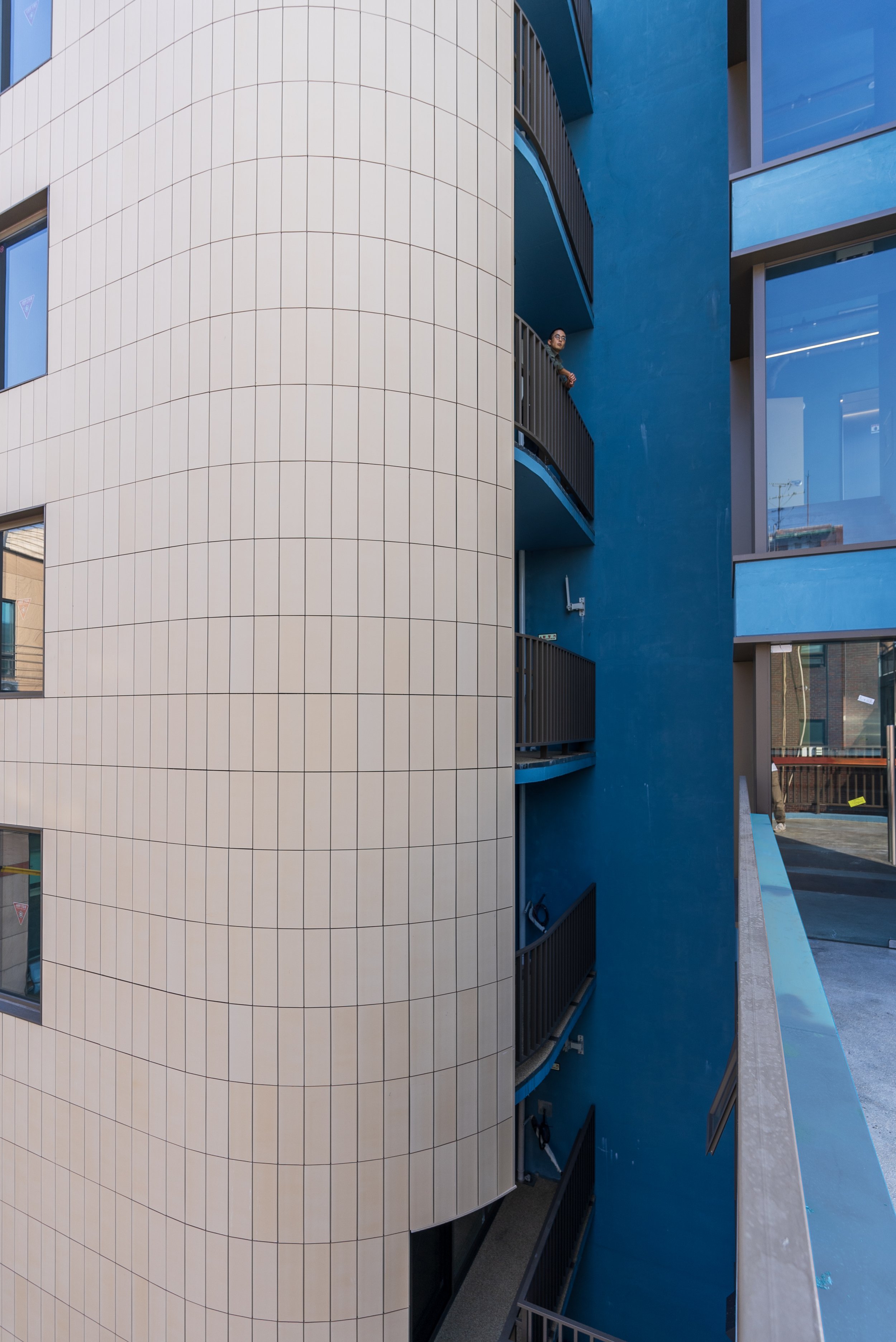 A man stands on a balcony of a modern blue building, looking out over the street. The building has balconies with black railings and large windows, with some reflection visible.