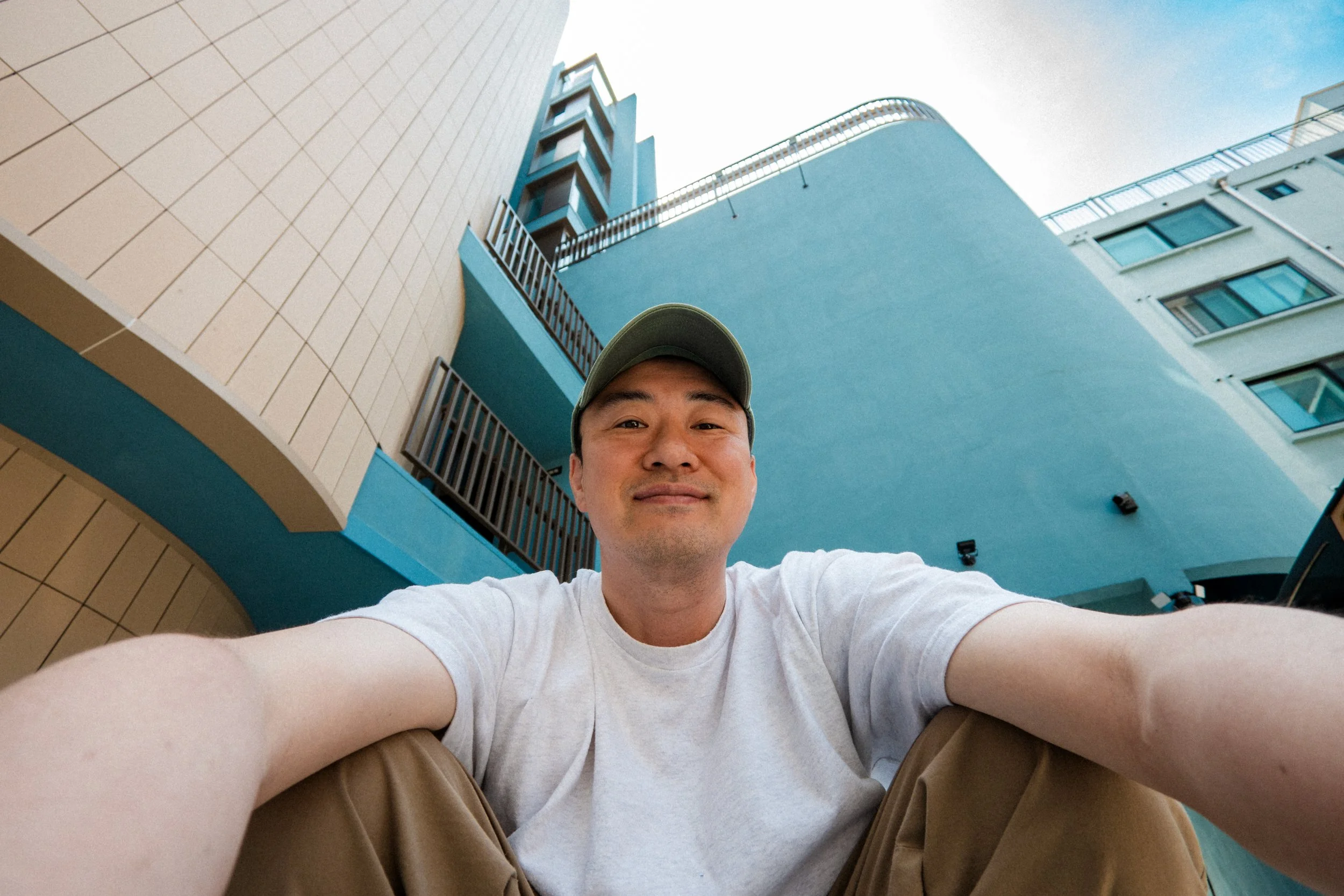 A young man taking a selfie with tall buildings in the background, smiling, wearing a baseball cap, a white t-shirt, and khaki pants.