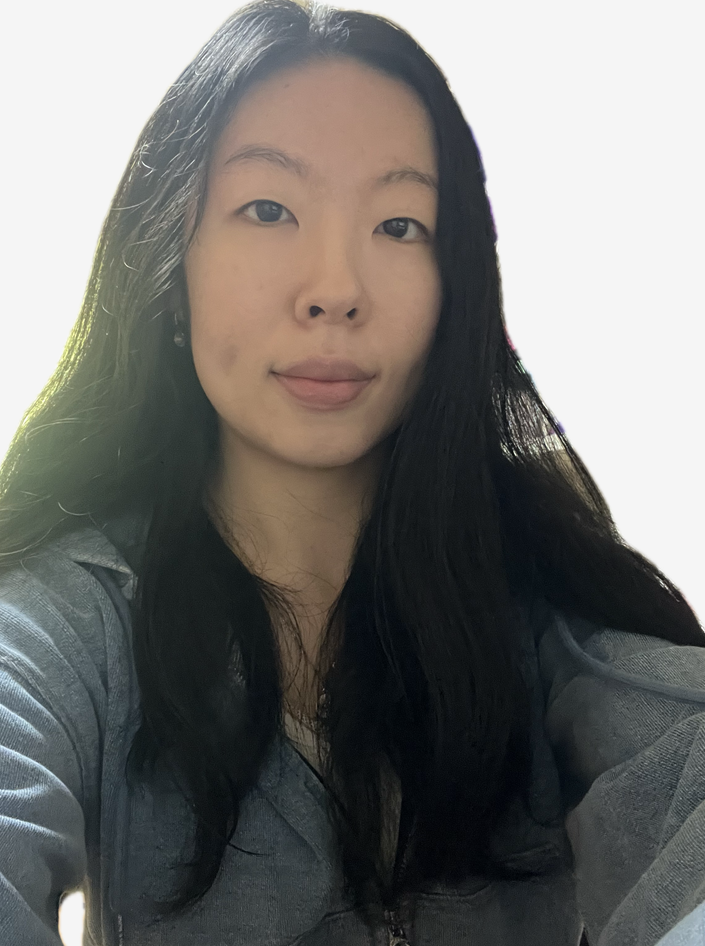 A young woman with long dark hair taking a selfie indoors, wearing a gray top.