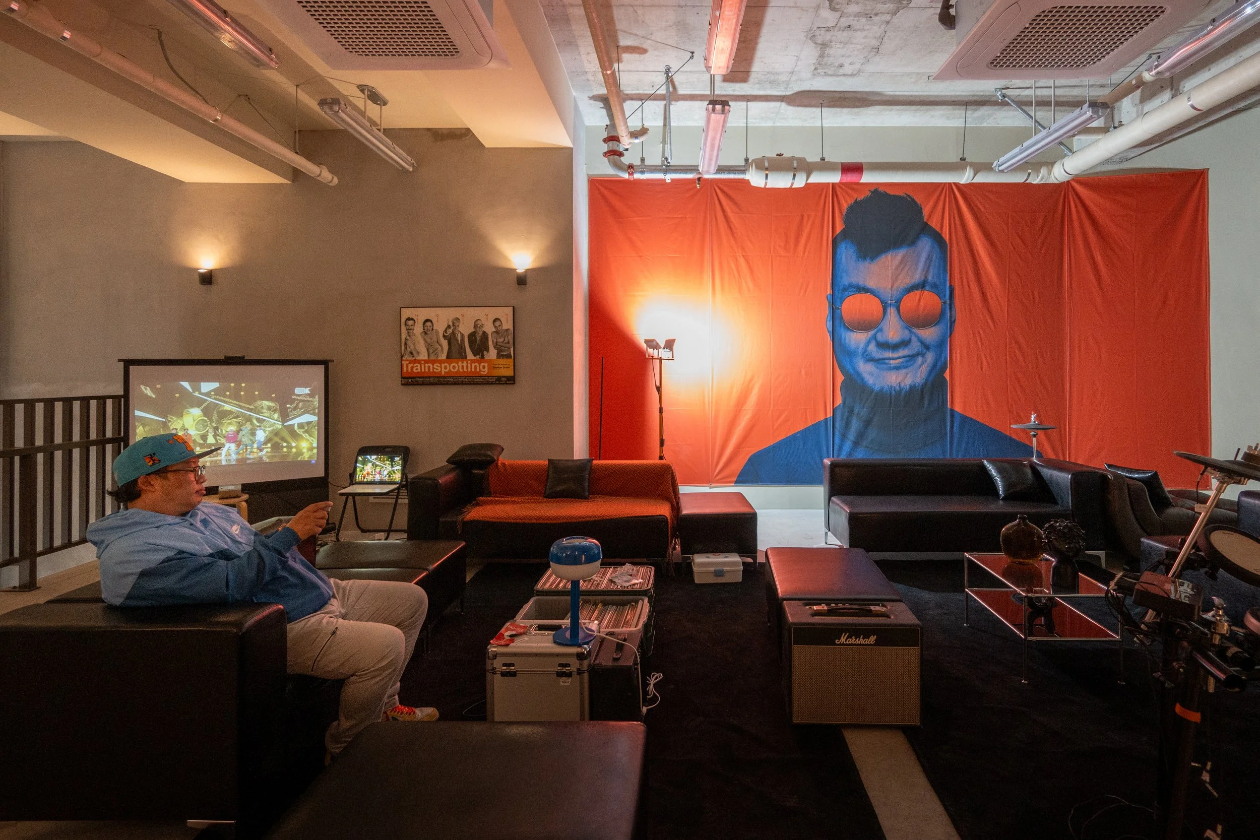 A dimly lit room with a large orange backdrop featuring a stylized portrait of a person with sunglasses, surrounded by modern furniture including sofas, a small table, and musical equipment, with a person sitting on a chair looking at a screen.