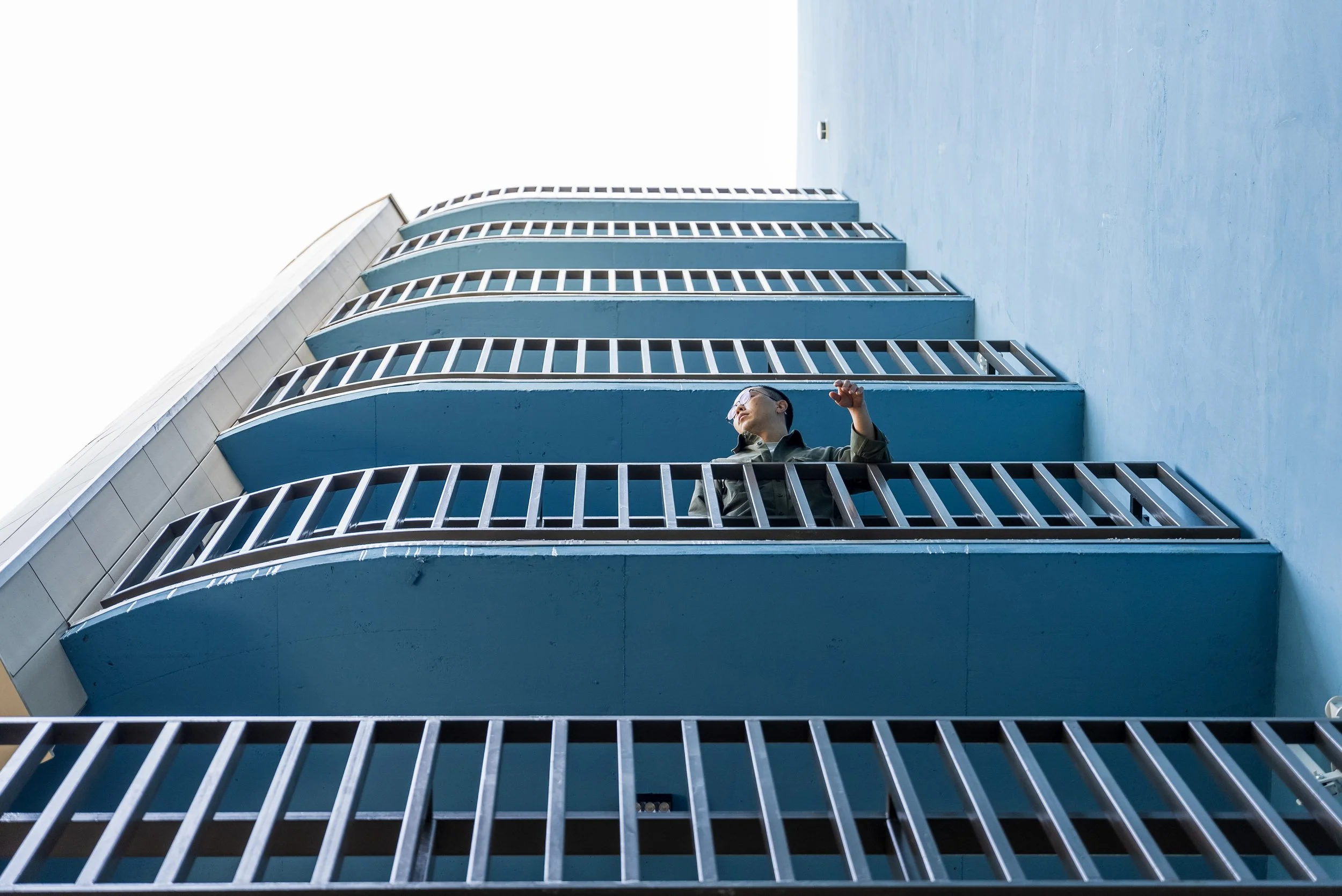 A woman standing on a blue balcony of a modern multi-story building, looking upwards with her hand raised.