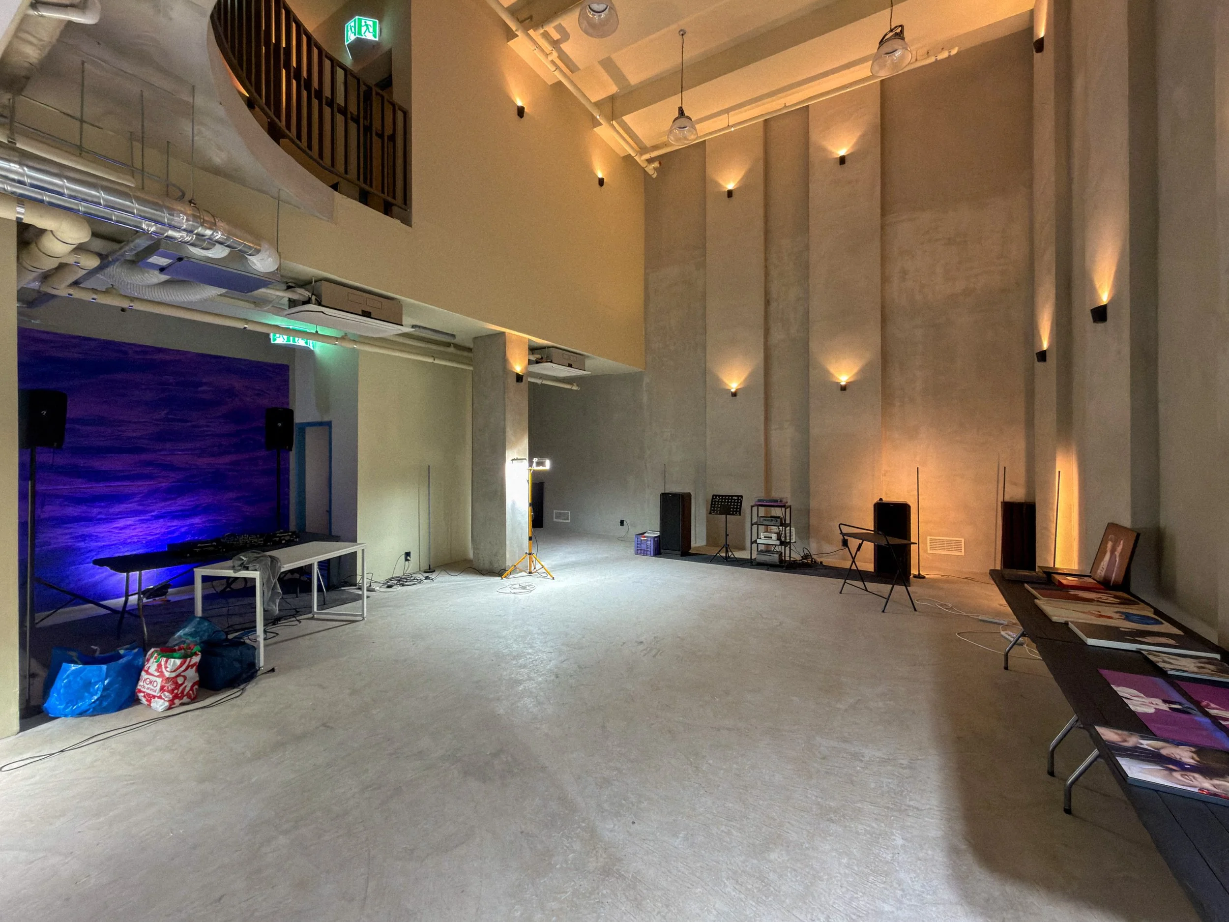 An empty event or performance space with a concrete wall, a small DJ setup with speakers and a table, and tables with magazines or art books. There are lighting fixtures on the wall and ceiling.