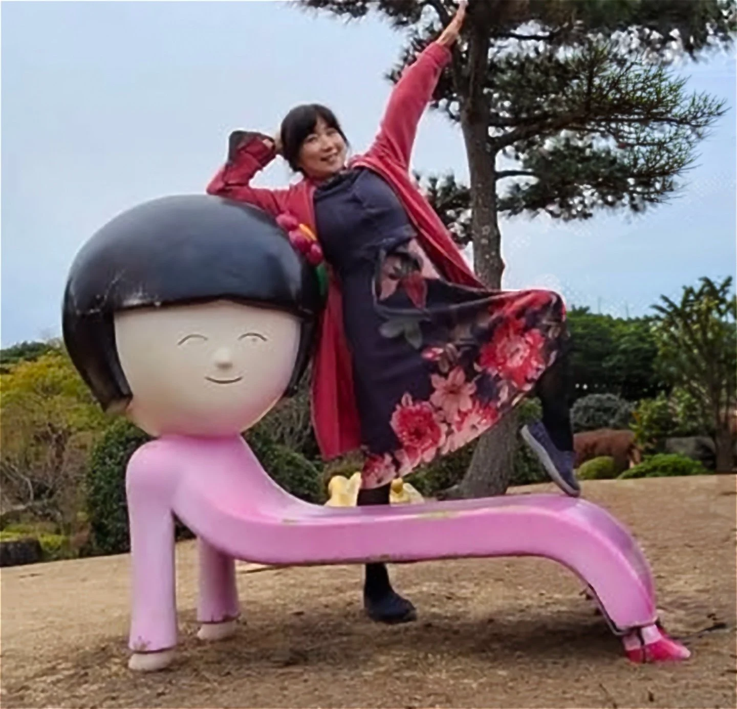 A woman with short dark hair, wearing a red jacket and floral dress, playfully poses on a large pink sculpture of a girl with a bob haircut, set outdoors among trees.