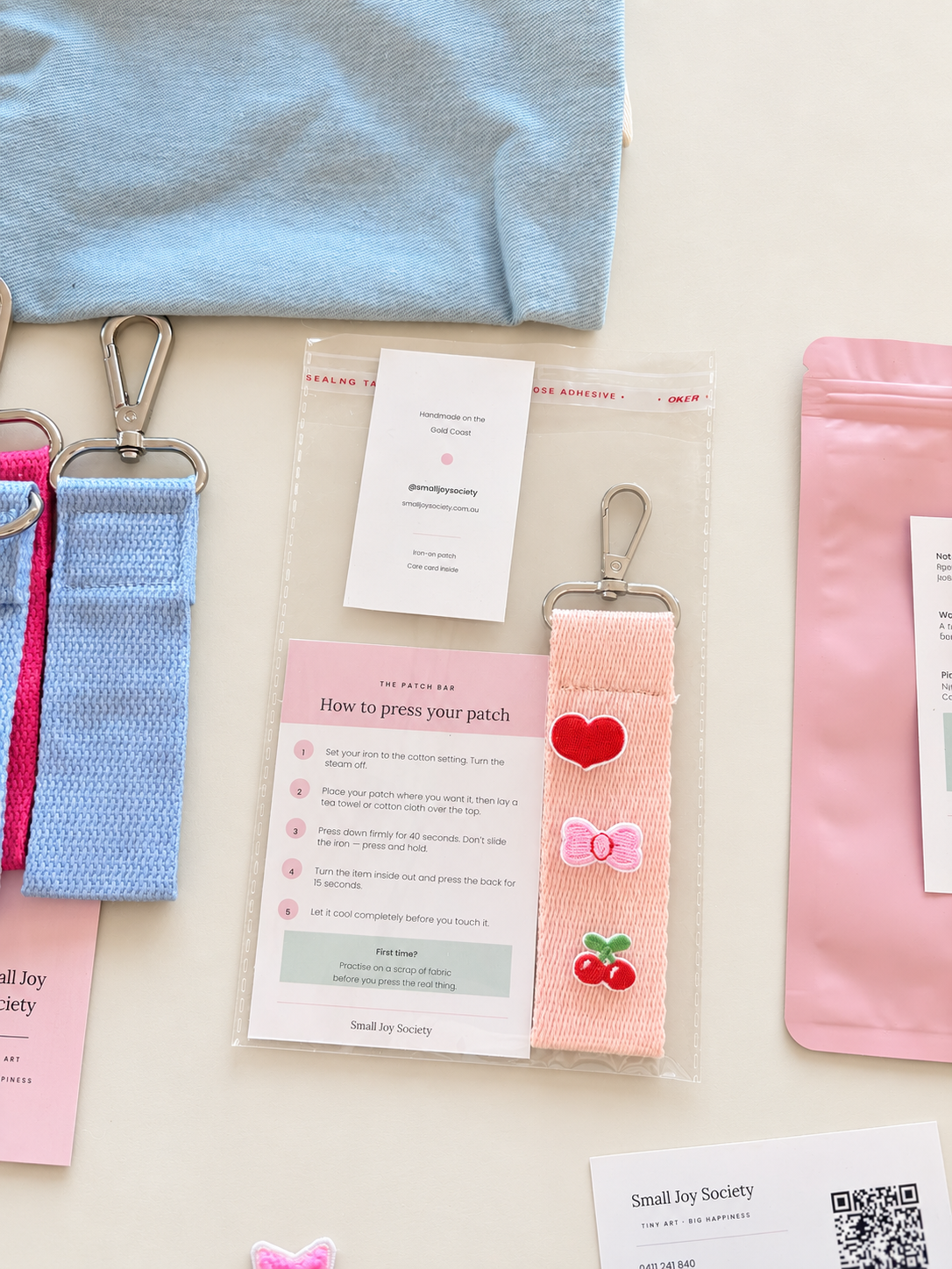 Small Joy Society Patch Bar wristlet with embroidered heart, bow and cherry patches beside the instruction card and gift packaging