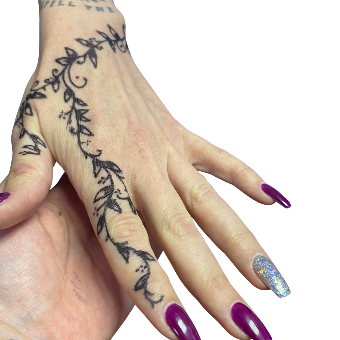 A hand with purple and glittery gel nails, adorned with a black ink floral tattoo extending from the fingers to the wrist.