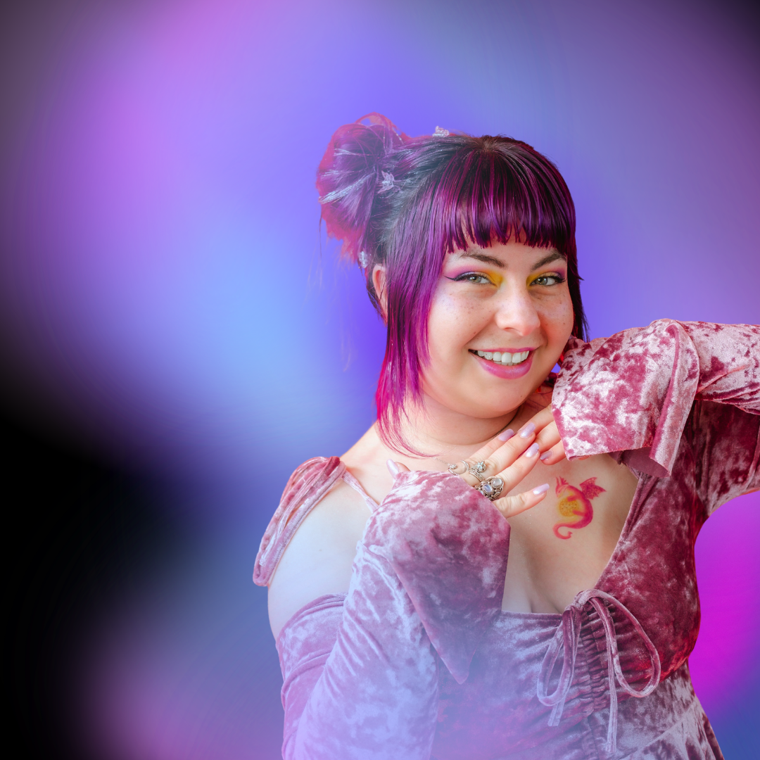 A woman with purple and pink hair in a messy bun, wearing a velvet mauve dress with shoulder cutouts, smiling, with colorful eye makeup and a dragon tattoo on her chest, posing with her hand near her face against a vibrant purple and blue gradient background.