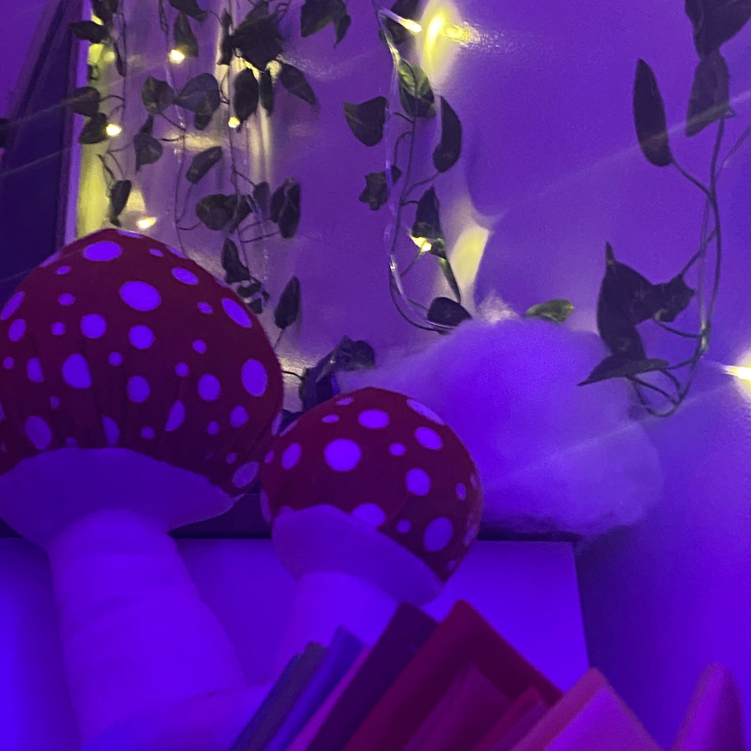 Decorative scene with two red mushrooms with white spots, green leaves, fairy lights, and fluffy white clouds in a dimly lit room.
