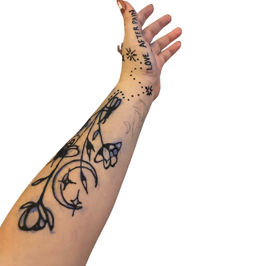 A person's forearm with a black tattoo of a floral pattern, butterflies, a crescent moon, and stars. The hand has words written on the fingers and wrist, including "LOVE" and "AFTER PAIN."
