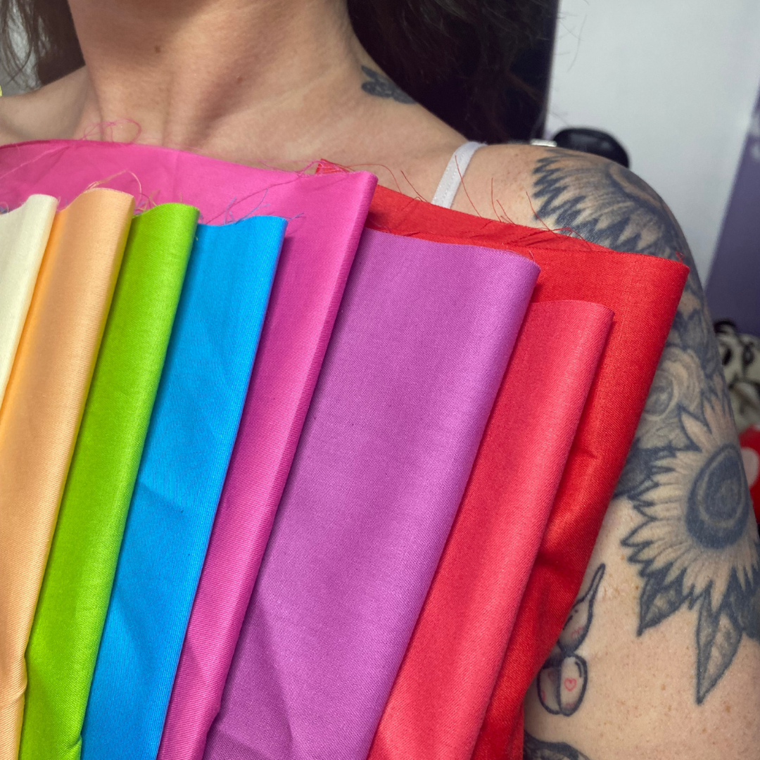 A person holding colorful fabric swatches in front of their chest and shoulder, with tattoos visible on their shoulder and arm.