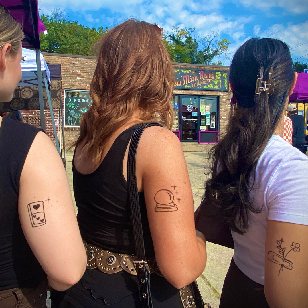 Three women standing outdoors at a market, with a storefront in the background called 'New Moon Rising Gifts & More'. The women have tattoos on their arms including a heart and stars on a phone, a crystal ball with stars, and a flower with a scroll lettering tattoo.