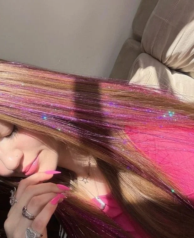 Close-up of a woman with long, straight hair with pink and purple highlights, wearing pink clothing and jewelry, touching her lips with her hand.