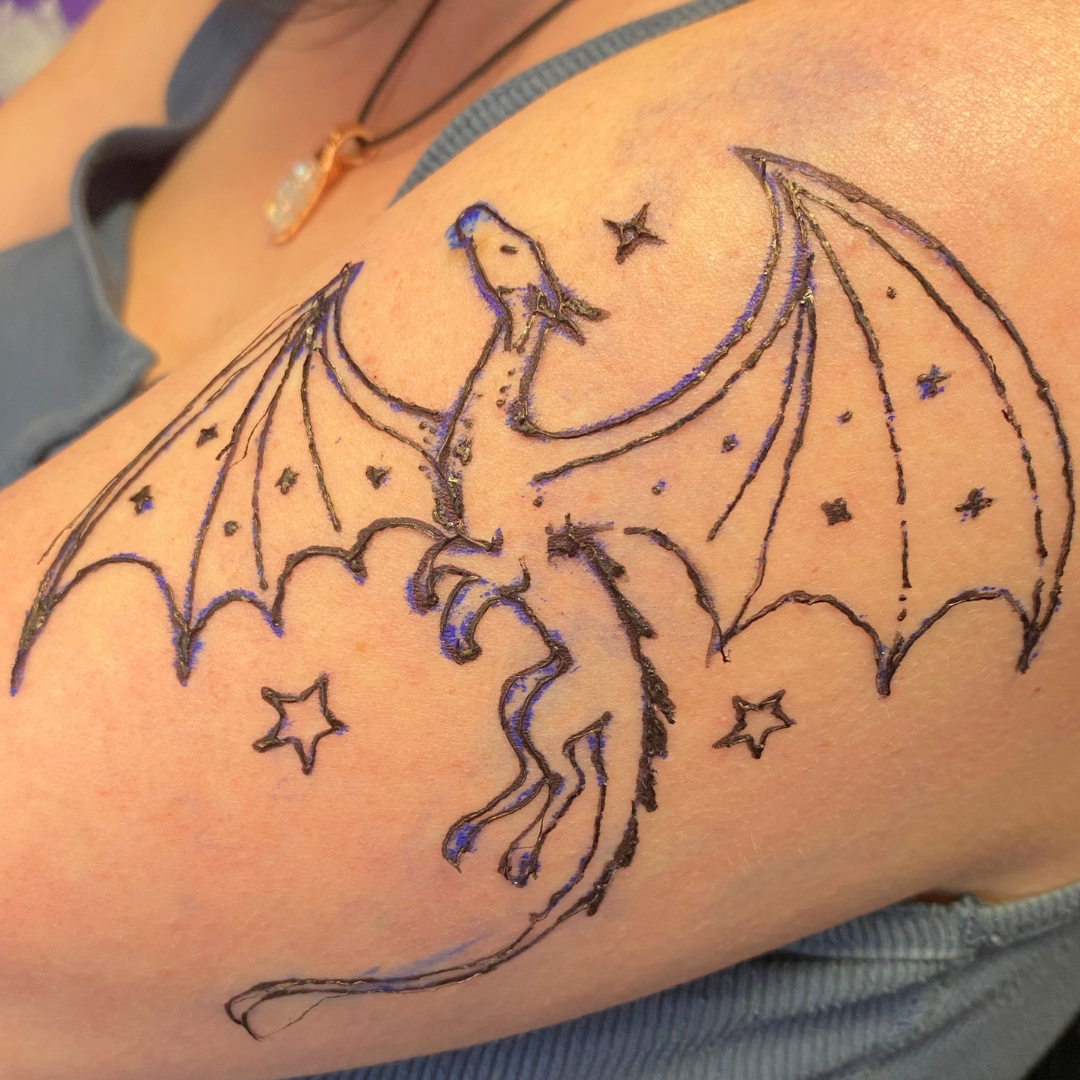 Tattoo of a dragon with wings, stars, and a crescent moon on a person's shoulder.
