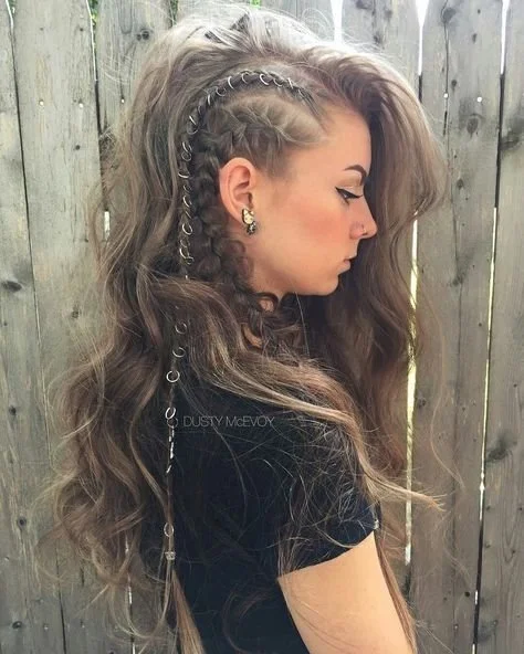 A woman with long, wavy brown hair featuring partially shaved design, styled with chains woven into her hair, standing in front of a wooden fence.