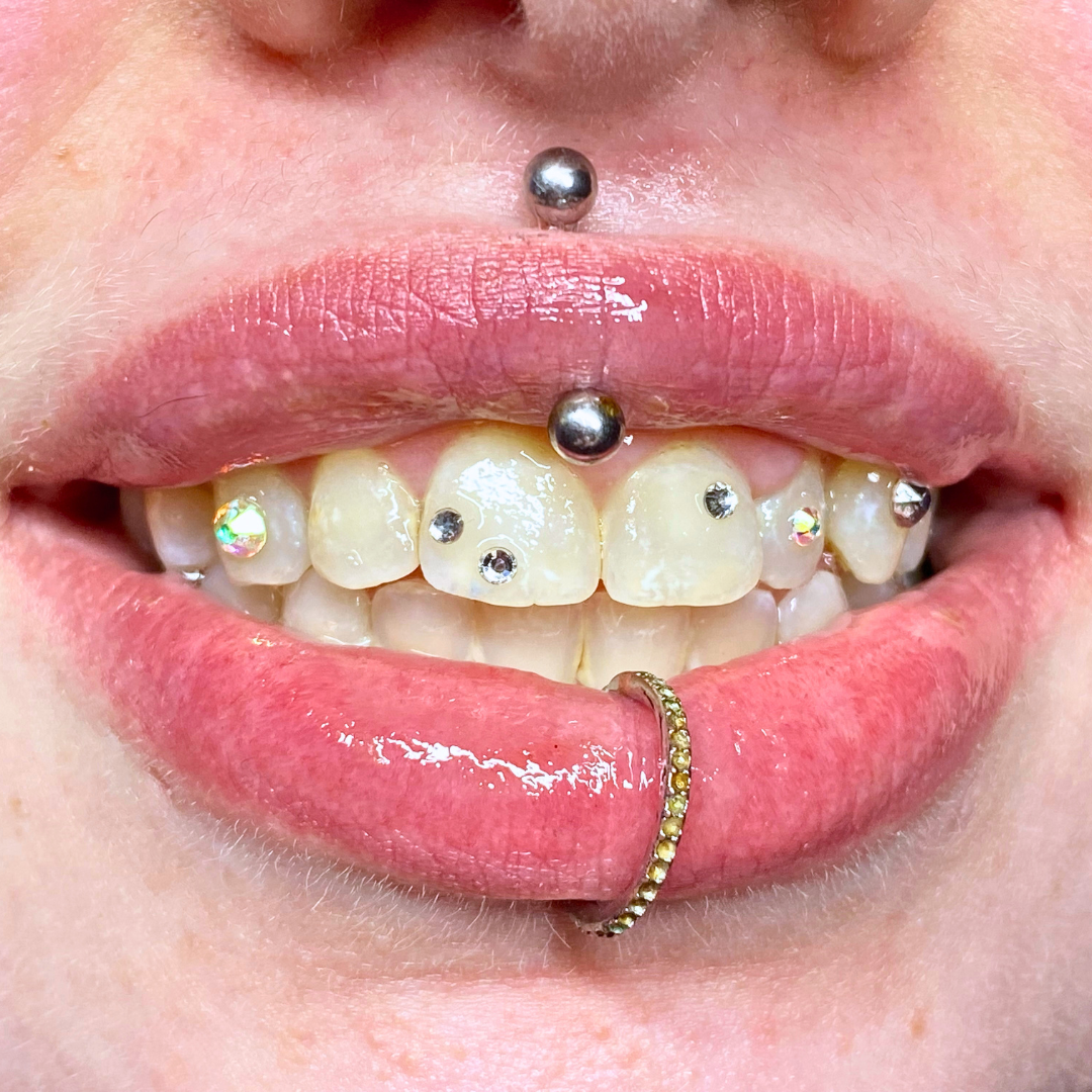 Close-up of a person's mouth showing multiple facial piercings, including a septum, mid-tongue, labret, and nose piercings, with decorated and pierced teeth.