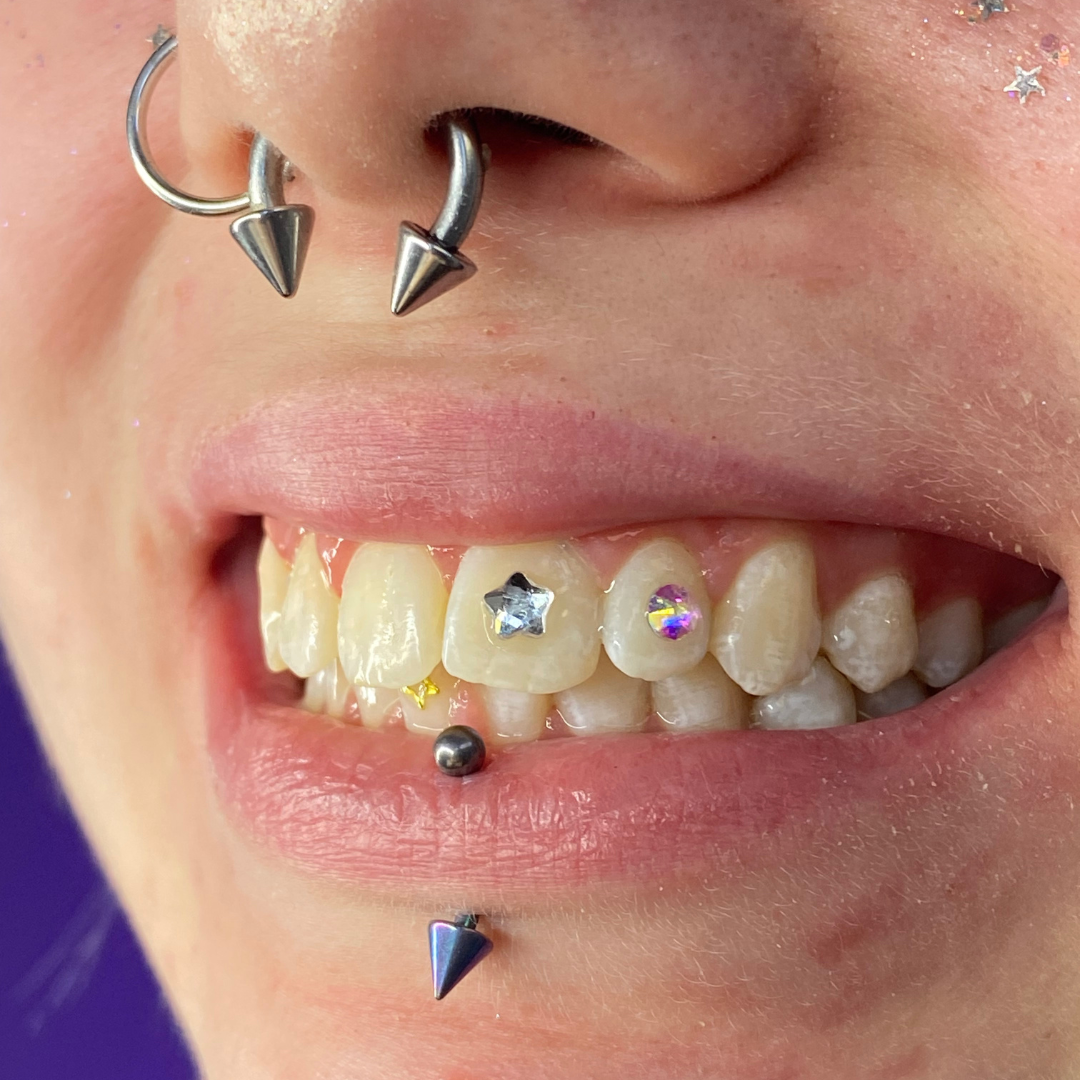 Close-up of a person's mouth showing multiple facial piercings, including two on the nose, a labret piercing below the lower lip, and a septum piercing. The person has several rhinestone decorations on their teeth, including a star-shaped one and oth