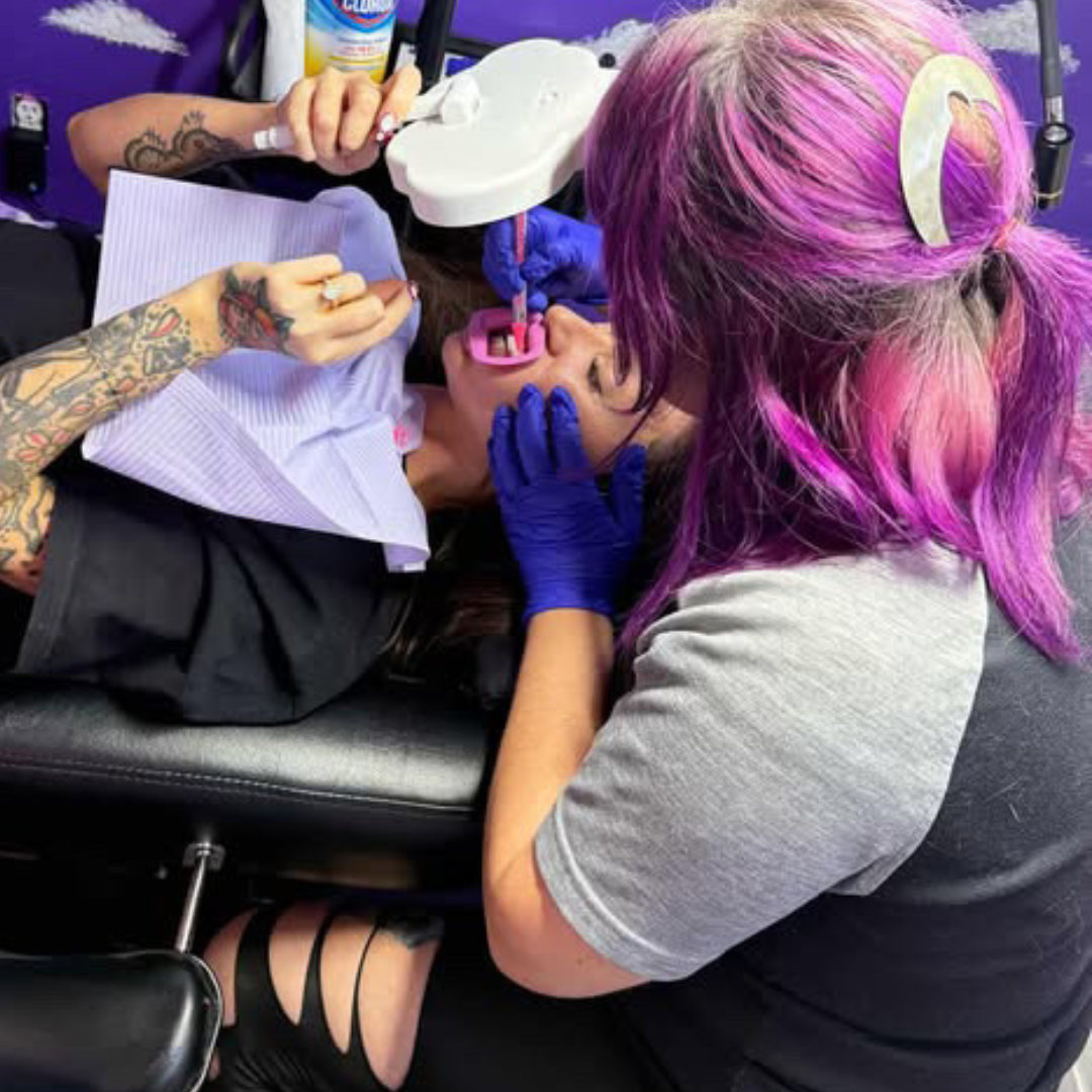 Tattooed woman with colorful hair, lying back in dental chair, receiving dental work from a technician with pink and purple hair, wearing blue gloves.