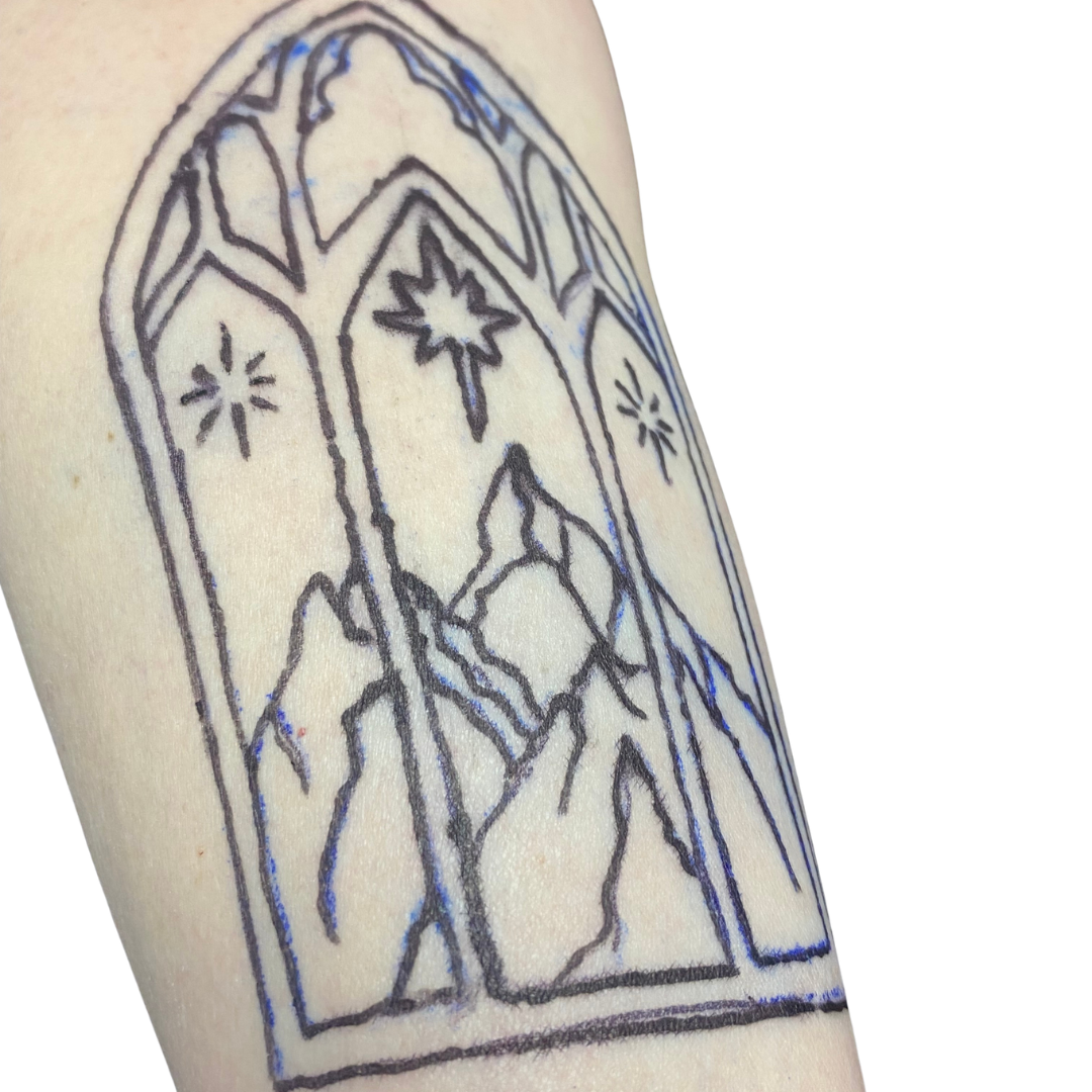Line art tattoo of a nativity scene with mountain background and stars, within an arched window frame.