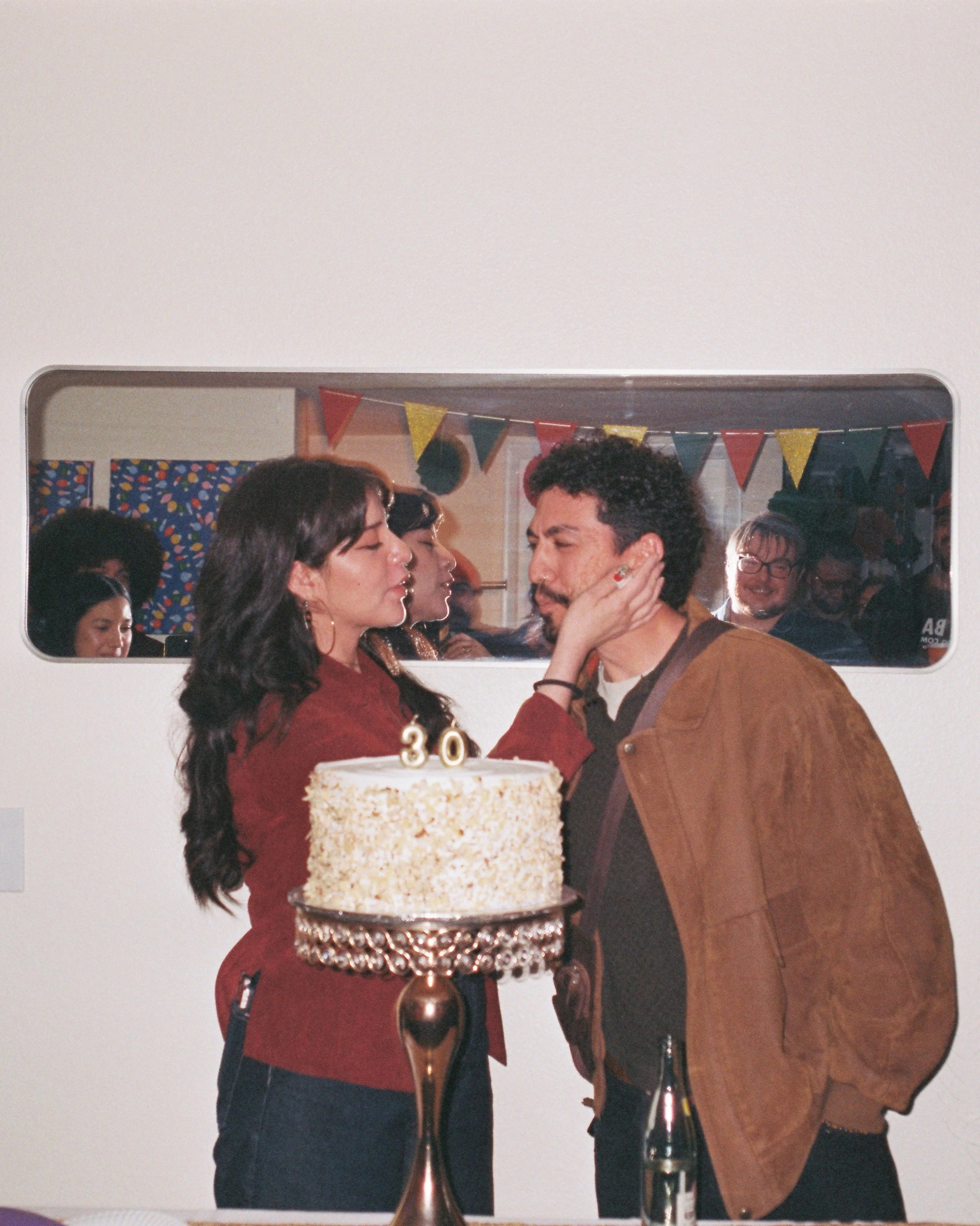 People gathered at a celebration, wedding or birthday, with a cake in the foreground, a couple embracing, and others in the background with balloons and decorations.