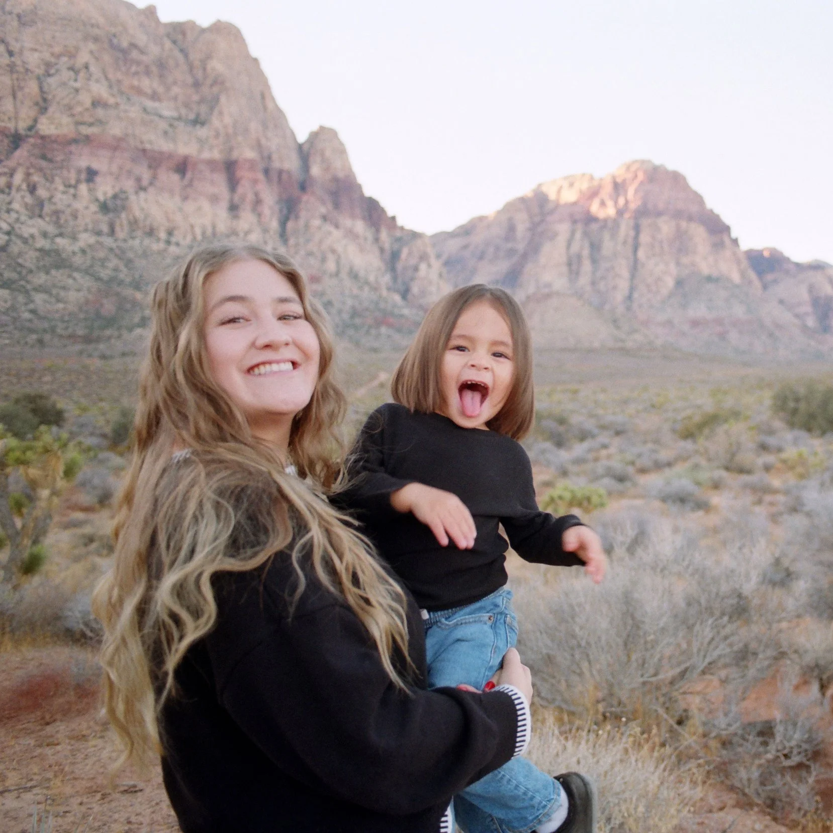 A smiling mother and son outdoors in a desert landscape with mountains in the background. The boy with brown hair is sticking her tongue out playfully.