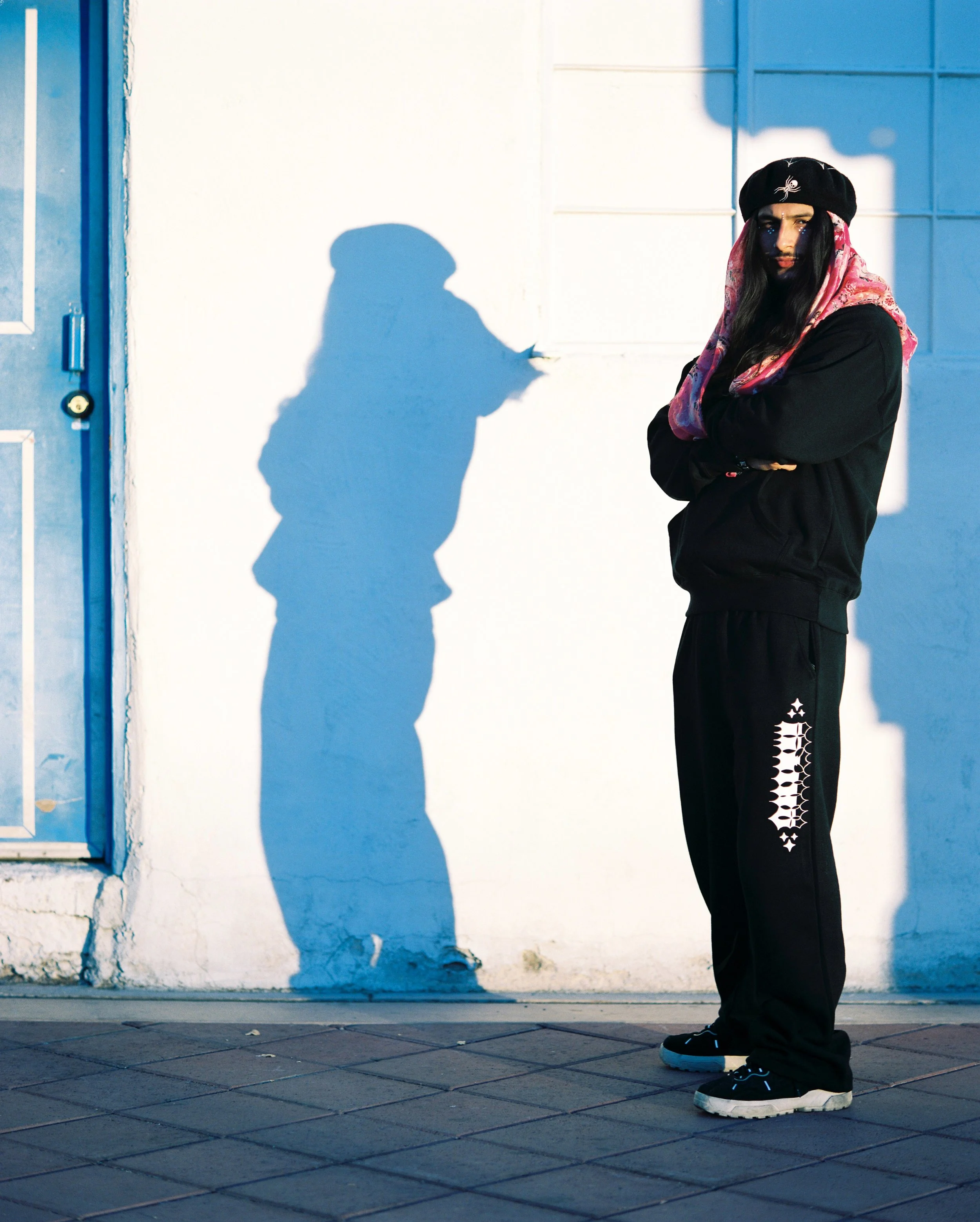 Person standing against a white wall with a blue door, wearing black sneakers, black pants with white decorative stitching, a black hoodie, a pink patterned hood, and a black beret with a white embroidered logo, casting a shadow on the wall.