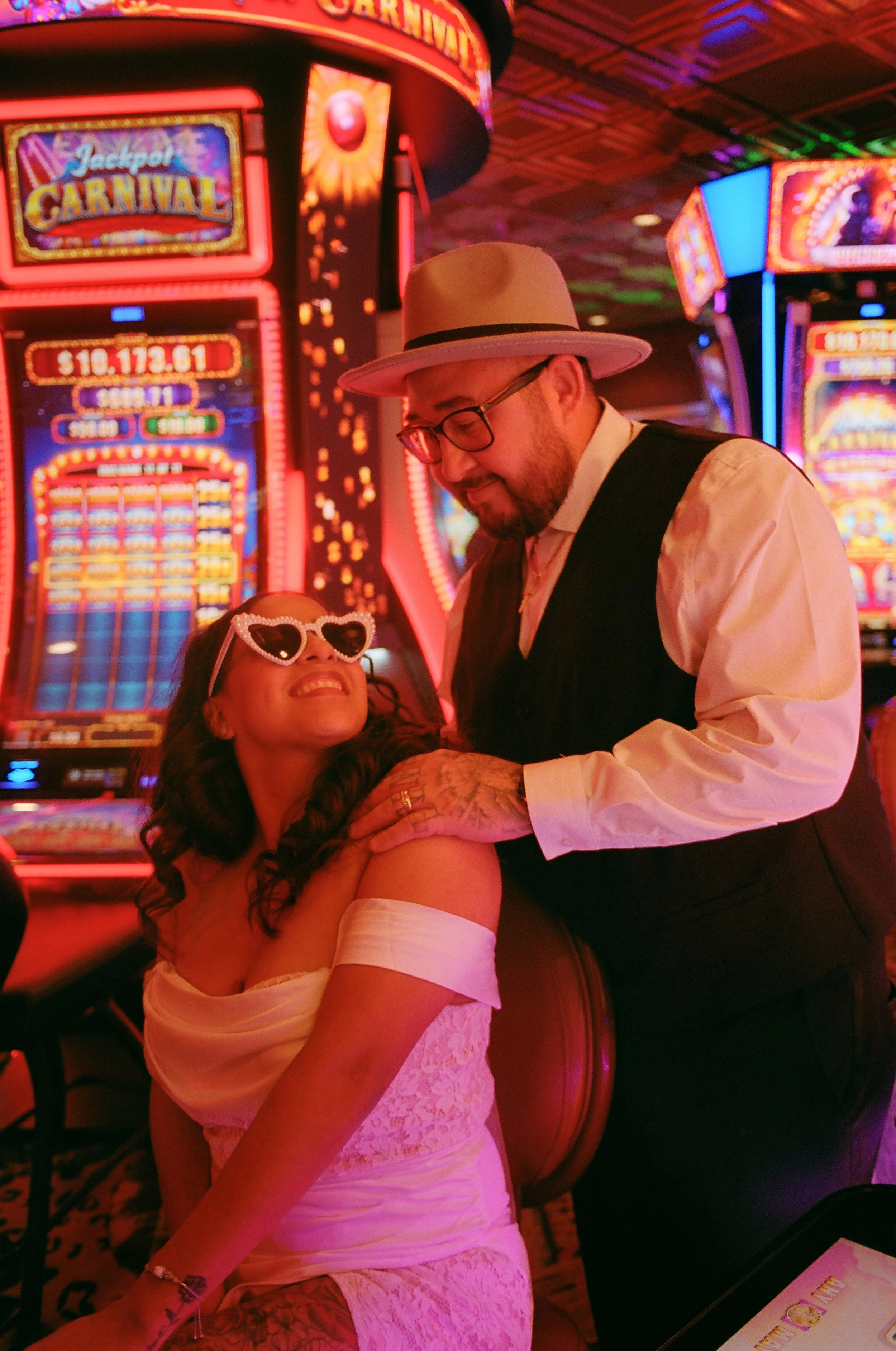 A woman in a white off-shoulder dress with heart-shaped sunglasses smiling as a man in a hat and glasses leans towards her, both in a casino with bright slot machines and colorful lights.